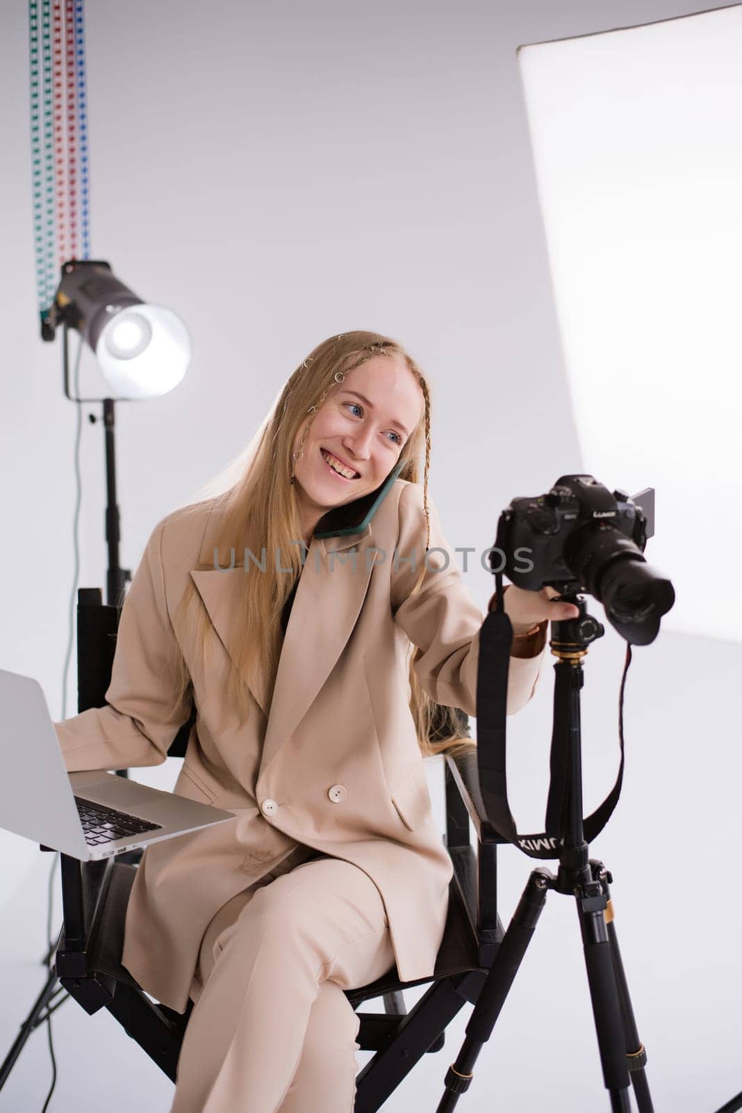 A woman producer videographer, a blonde with a camera, smartphone and laptop MacBook happy talking on the phone and smiling in the photo studio. Wearing a formal nude pantsuit on a white background