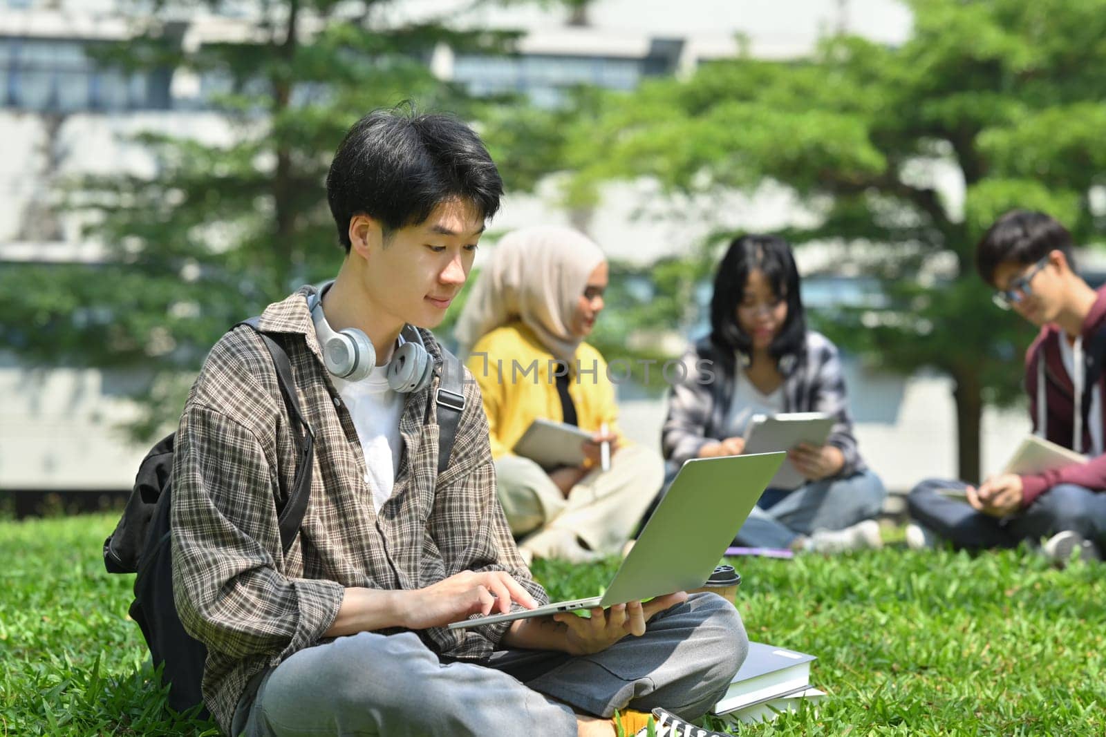Young asian man student using laptop on green grass in front of university building. Education, technology and lifestyle concept.