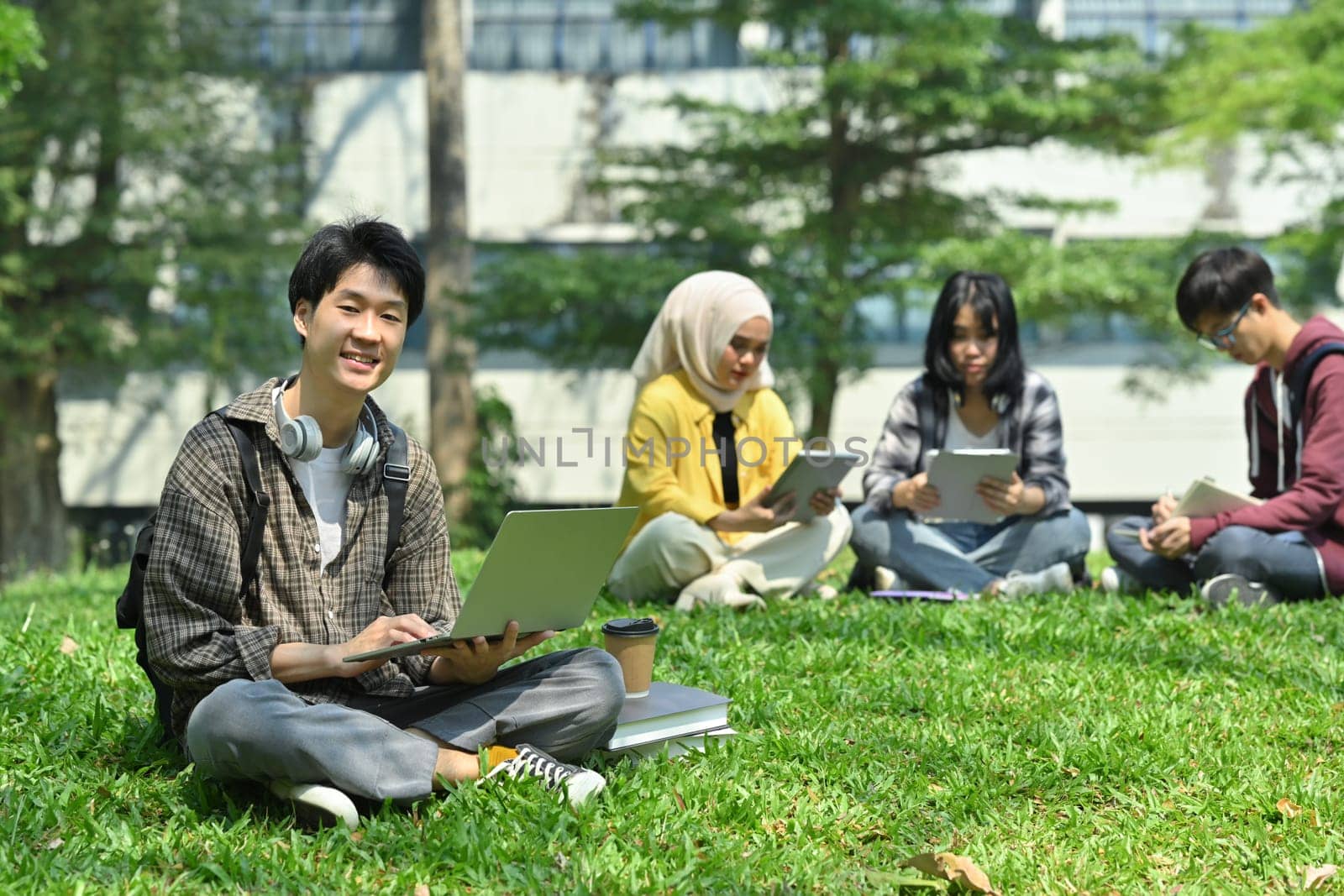 Young asian man student using laptop on green grass in front of university building. Education, technology and lifestyle concept by prathanchorruangsak