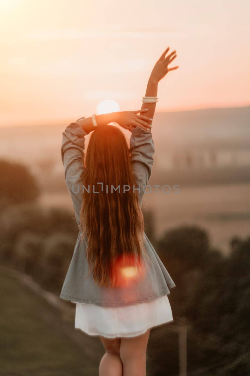 Happy woman standing with her back on the sunset in nature in summer with open hands. Romantic beautiful bride in white boho dress posing with mountains on sunset by panophotograph