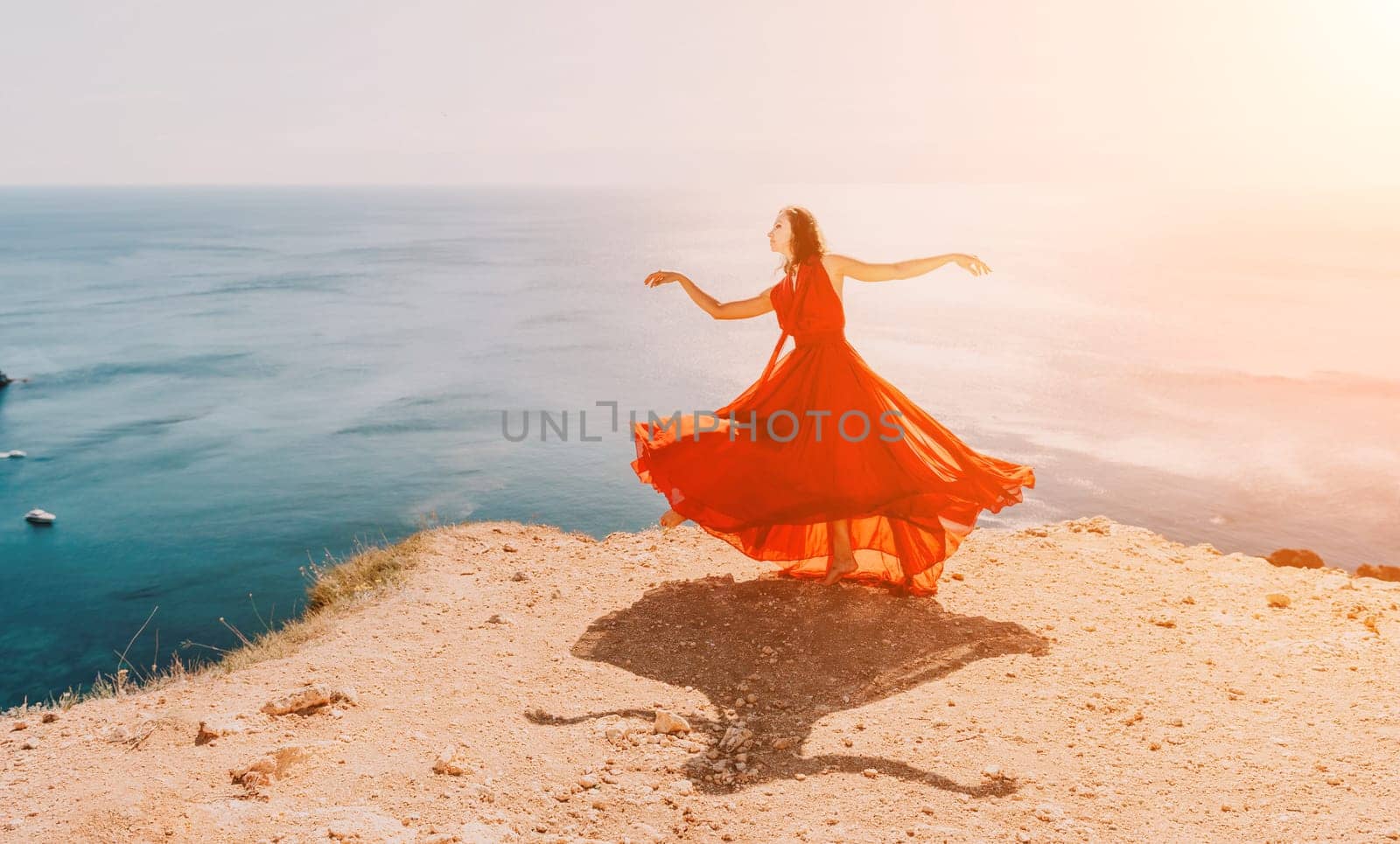 Woman in red dress on sea. Side view a Young beautiful sensual woman in a red long dress posing on a rock high above the sea on sunset. Girl on the nature on blue sky background. Fashion photo. by panophotograph