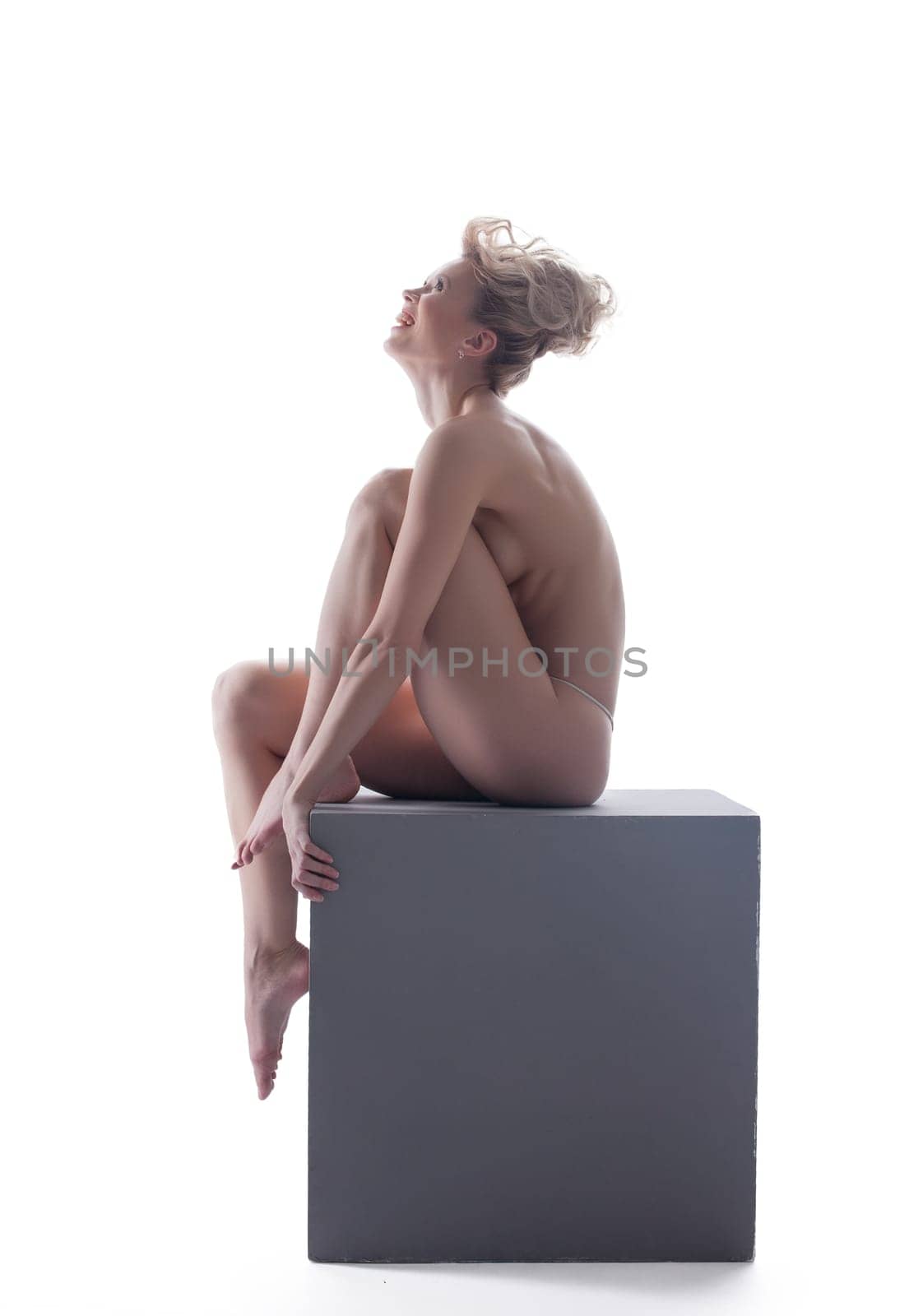 Merry young blonde smiling while sitting on cube, isolated over white background