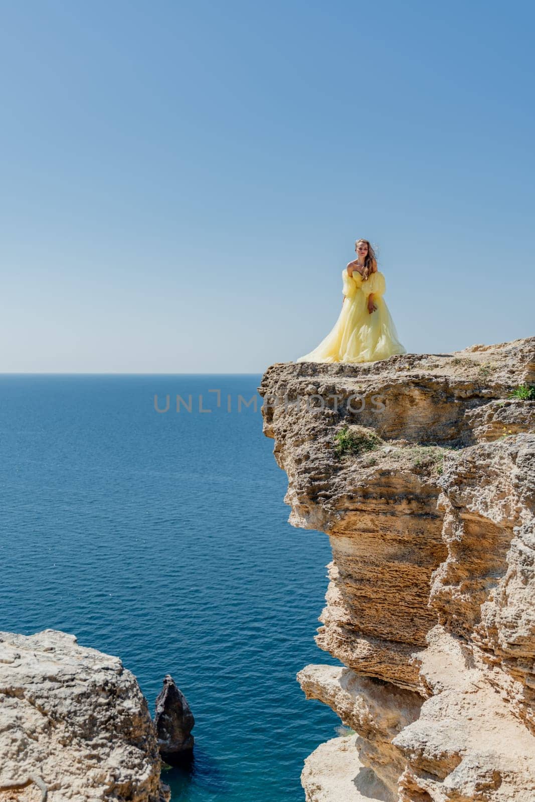 Woman in a yellow dress on the sea. Side view Young beautiful sensual woman in yellow long dress posing on a rock high above the sea at sunset. Girl in nature against the blue sky by Matiunina