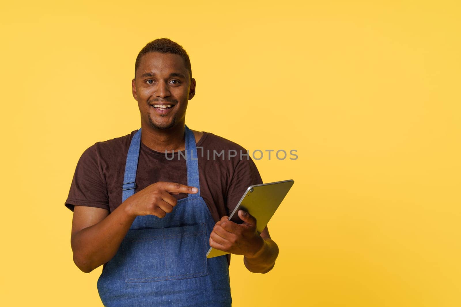 Young, talented African American chef search for the perfect recipe on his trusty tablet. Clad in a stylish blue apron isolated on yellow background, he symbolizes the fusion of modern technology and traditional gastronomy. High quality photo