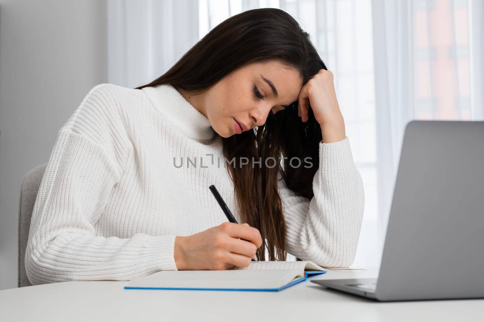 Portrait of a thoughtful student makes notes during lectures online while sitting with a laptop at home.