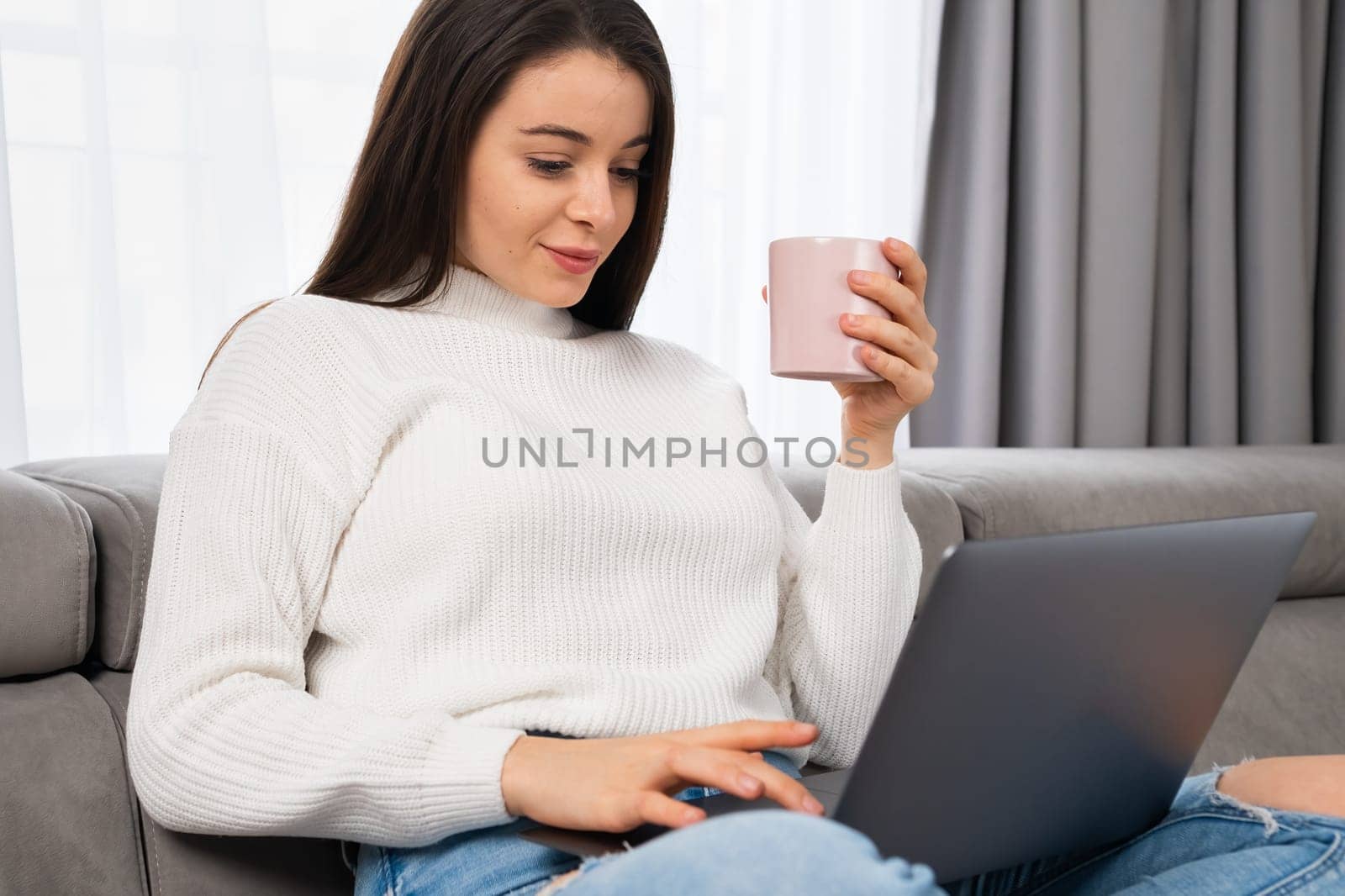 Woman relaxing with a cup of tea at home using a laptop in a modern apartment.