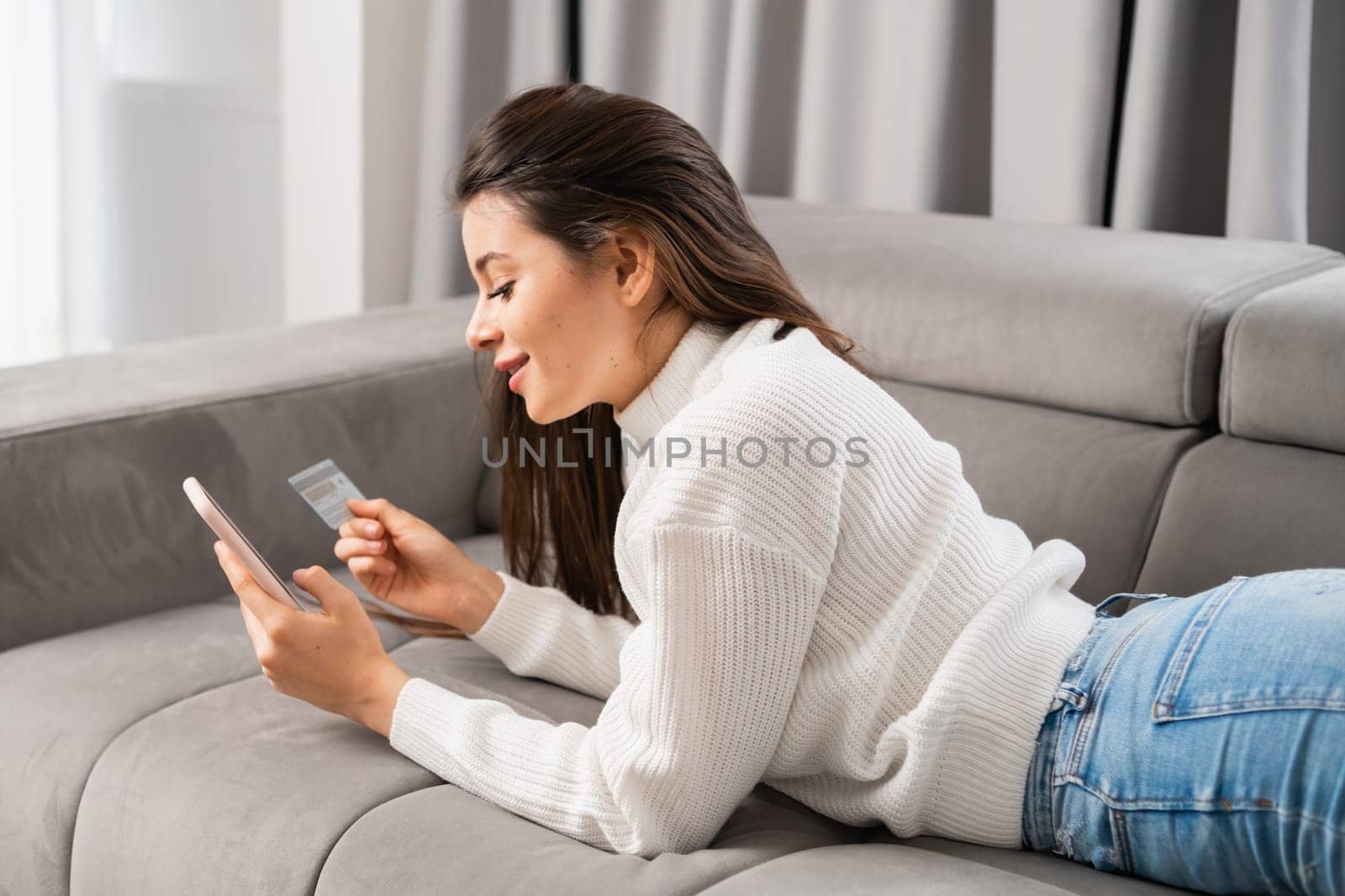 A beautiful young woman lying on the sofa makes purchases via the internet and pays by card