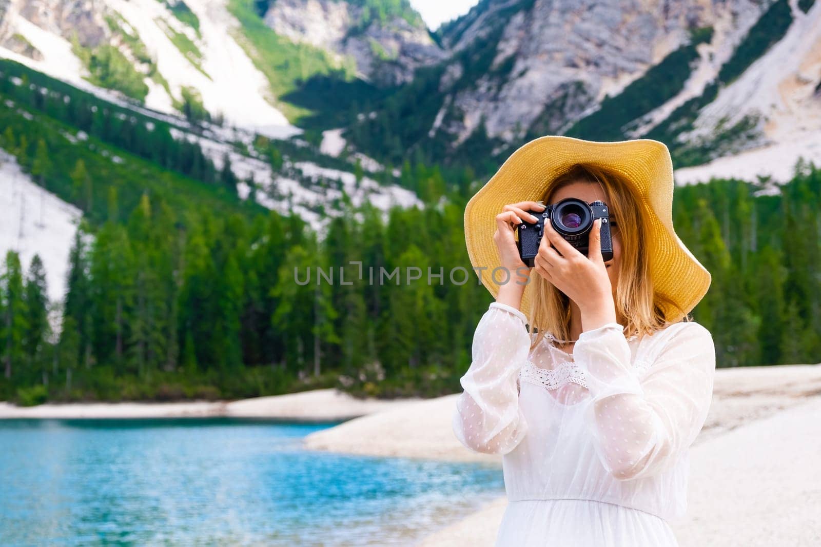 A beautiful blond hair woman photographer in a hat and white dress takes a photo on the shores of lake Braies with Dolomites Alps on the background with copy space. Travel agency advertising concept