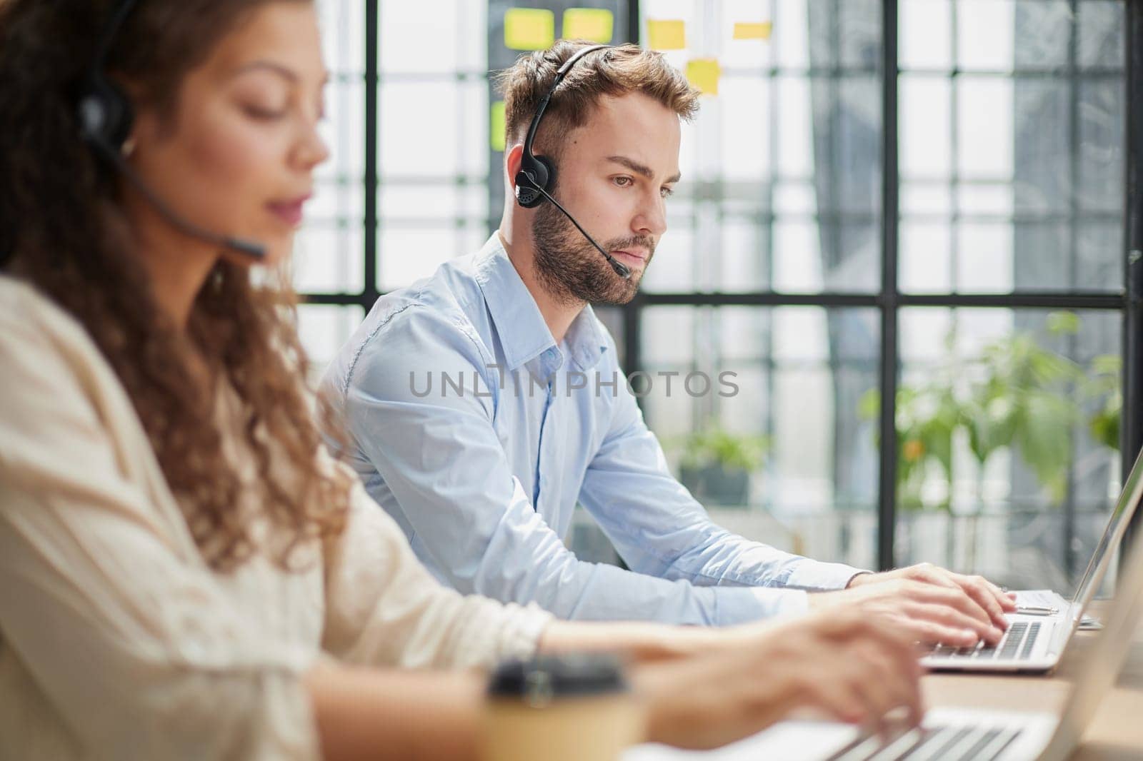 male call-center operator with headphones sitting at modern office, consulting online information in a laptop, looking up information in a file in order to be of assistance to the client.