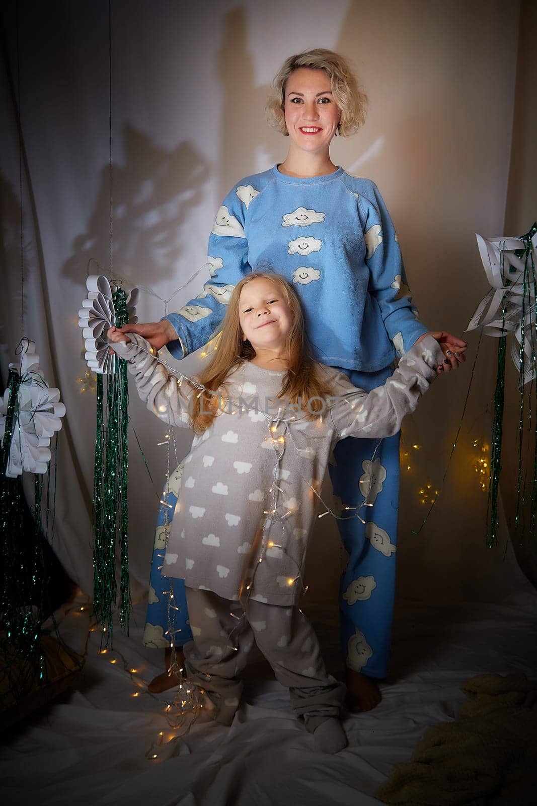 Cute mother and daughter in pajamas having fun in the room with Christmas garlands and white background. The tradition of decorating the house for holidays. Happy childhood and motherhood