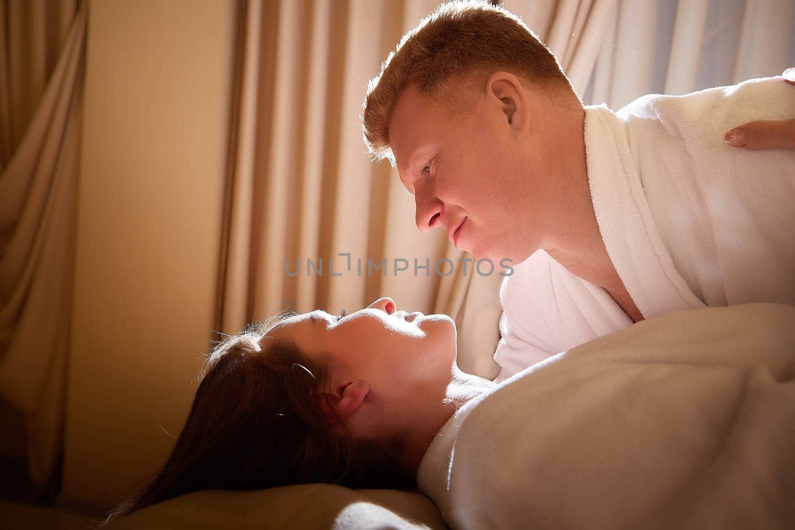 An adult couple of newlyweds in in white robes in a hotel room after the wedding. A guy and a girl, a man and a woman in beautiful room. The concept of hugs, love and care