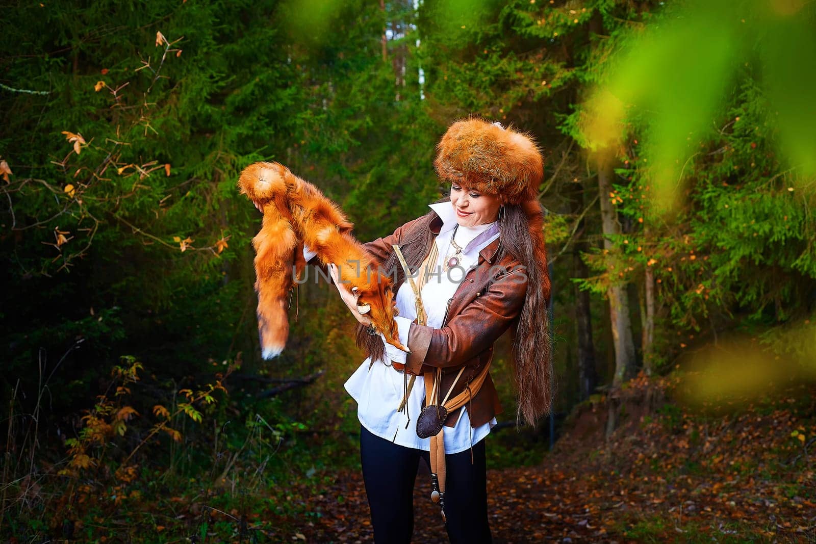 Girl in a leather jacket, a big red fox fur hat and with the skin of a fox killed on the hunt in the forest in autumn. A female model poses as fabulous royal huntress on nature hunt at photo shoot