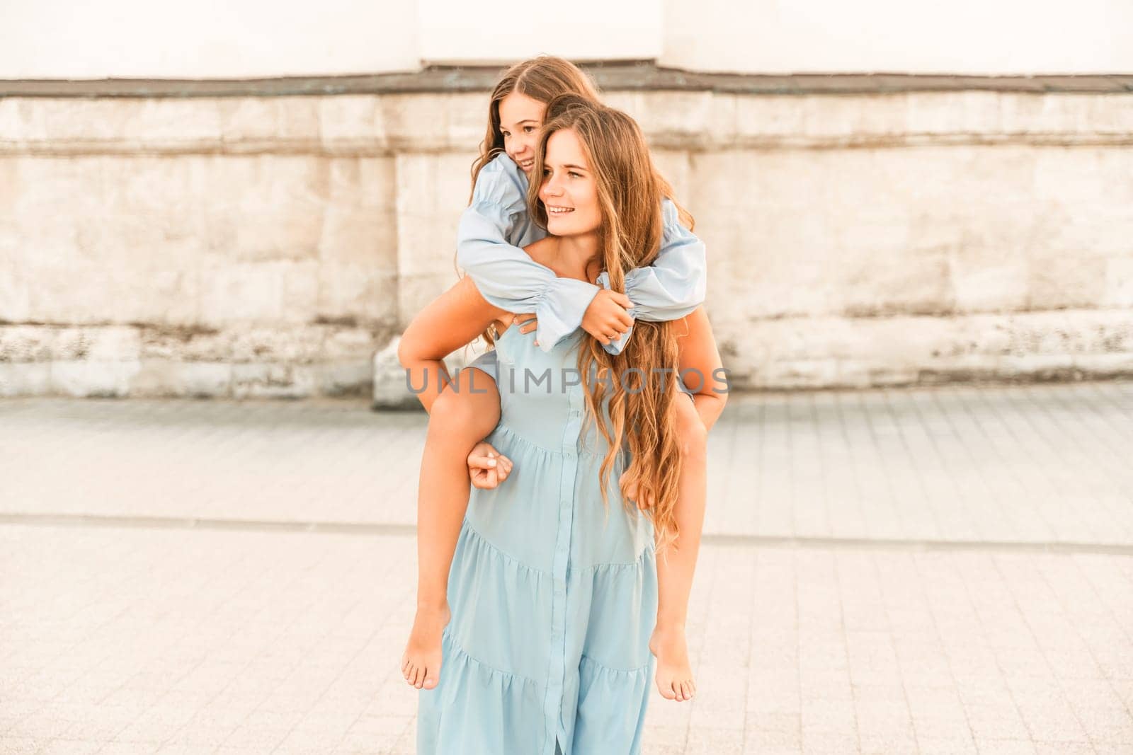 Mother of the daughter walks playing. Mother holds the girl on her back, holding her legs, and her daughter hugs her by the shoulders. Dressed in blue dresses. by Matiunina
