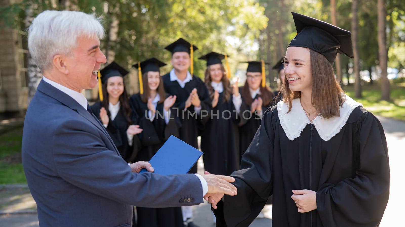 The teacher shakes hands with the student and presents the diploma outdoors. A group of university graduates