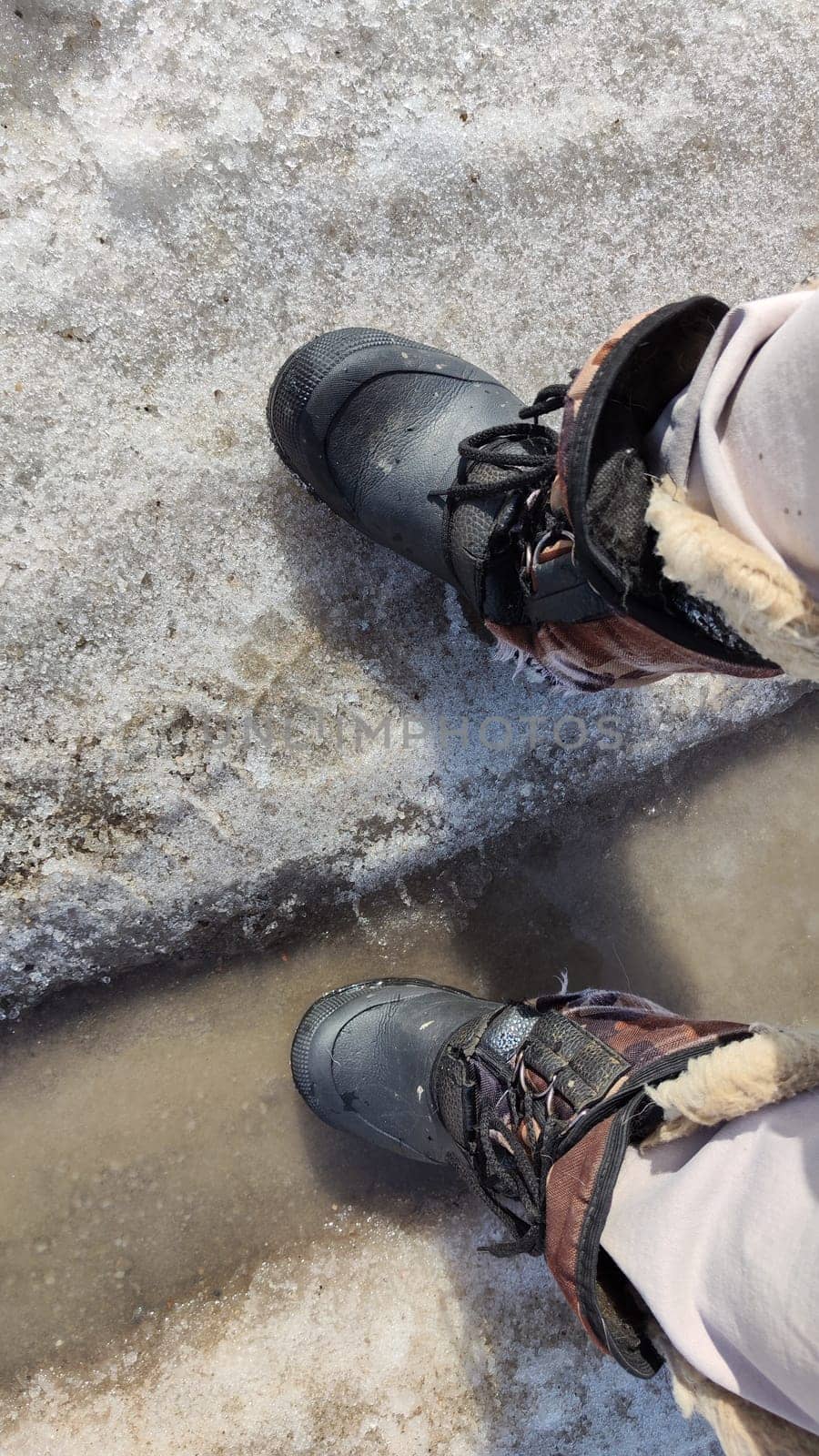 Feet of Hunter or fisherman in big warm boots on winter day on snow. Top view. Fisherman on the ice of a river, lake, reservoir on spring day with melting ice. Dangerous fishing in autumn