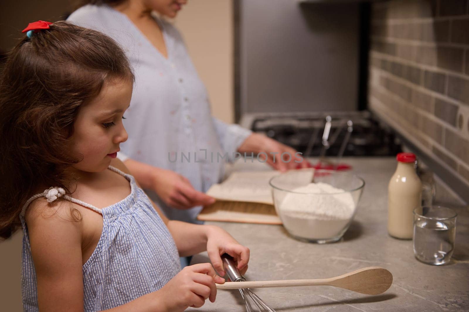 Beautiful child, adorable little girl standing at kitchen counter top, holding a wooden spoon on the blurred background of her mom, searching a recipe on recipe book for preparing pancake dough