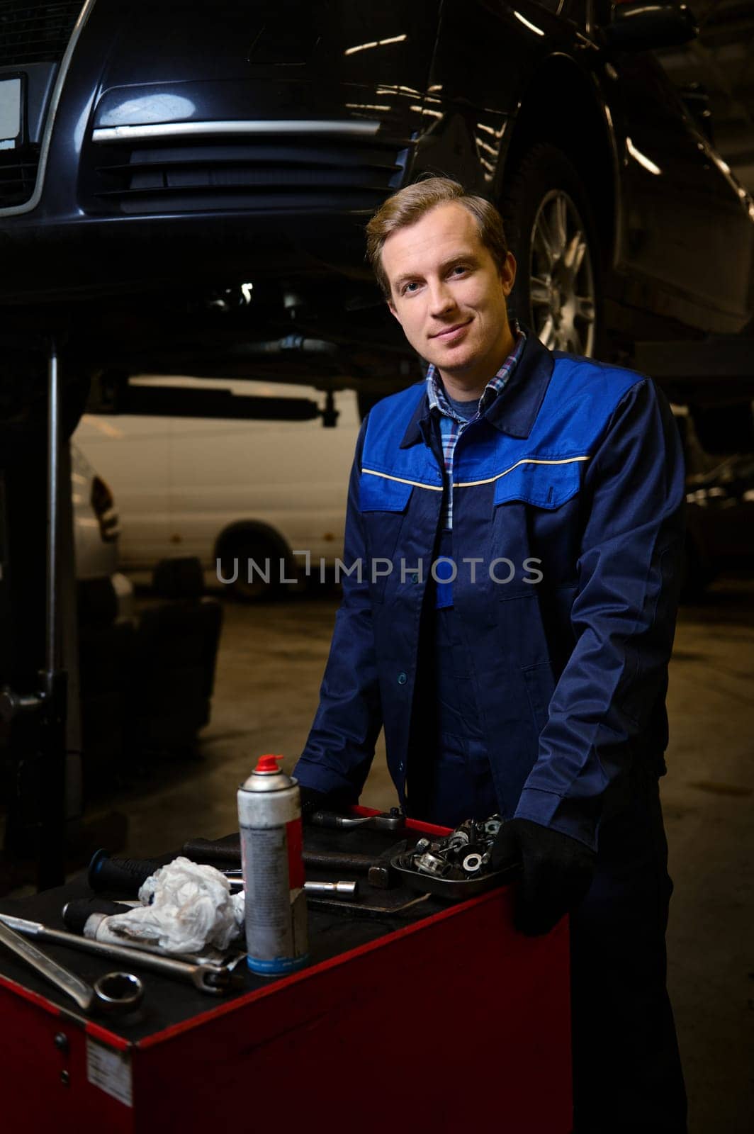 Professional portrait of a handsome Caucasian man, auto mechanic, technician, car engineer standing near a box with set of tools for repairing a lifted automobile on a hoist in the repair shop garage