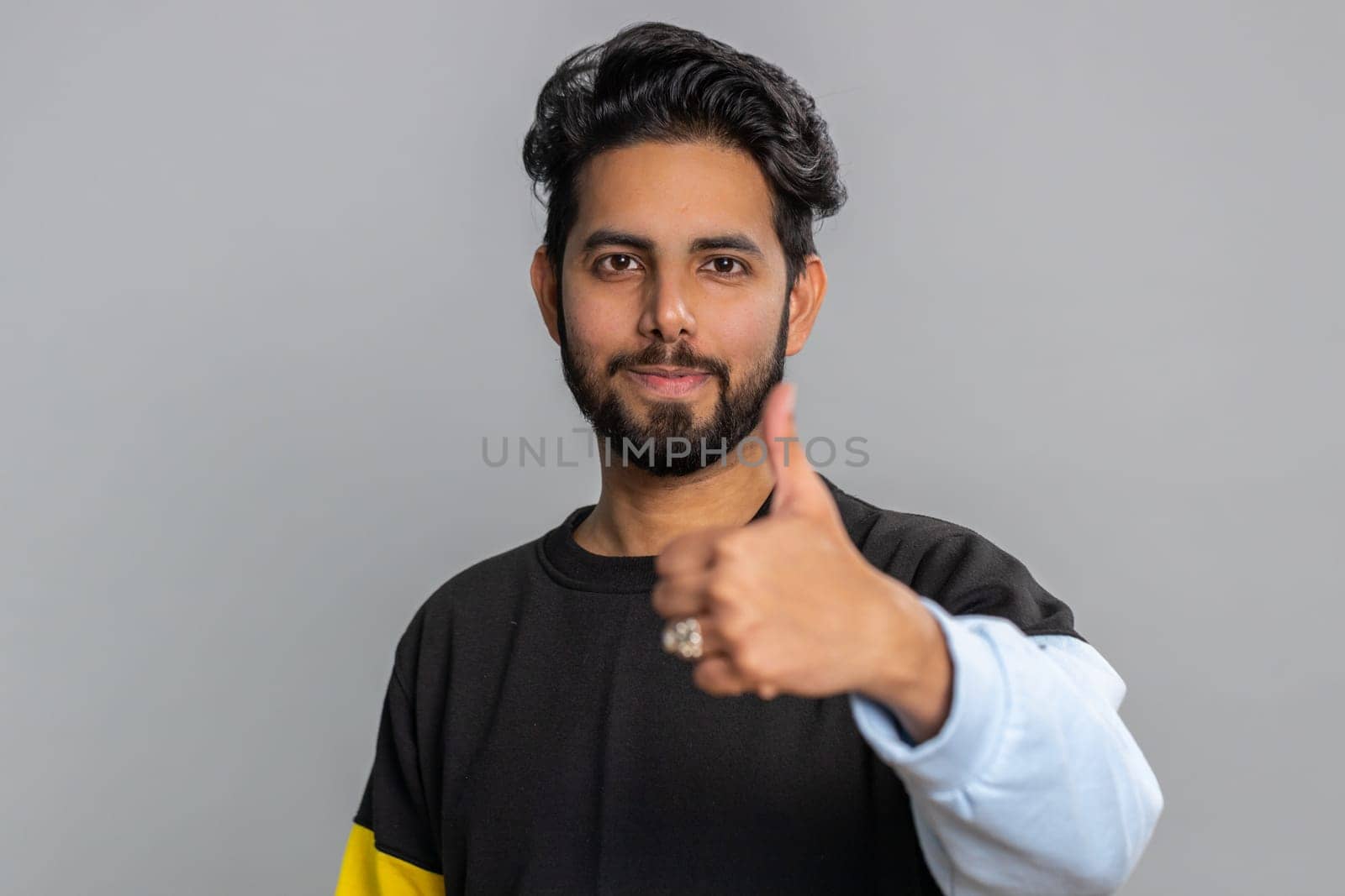 Like. Indian young man in sweater raises thumbs up agrees with something, gives positive reply recommends advertisement likes good. Handsome bearded hindu guy isolated alone on gray studio background