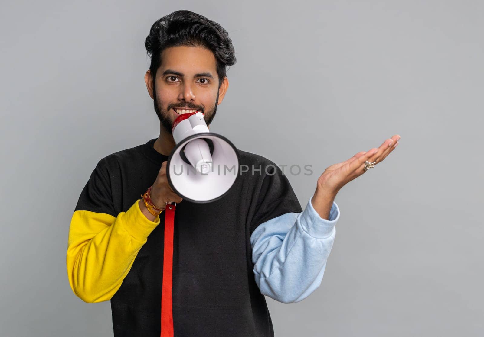 Indian man talking with megaphone, proclaiming news, loudly announcing advertisement, warning using loudspeaker to shout speech, pointing empty place. Copy-space. Handsome guy on gray wall background