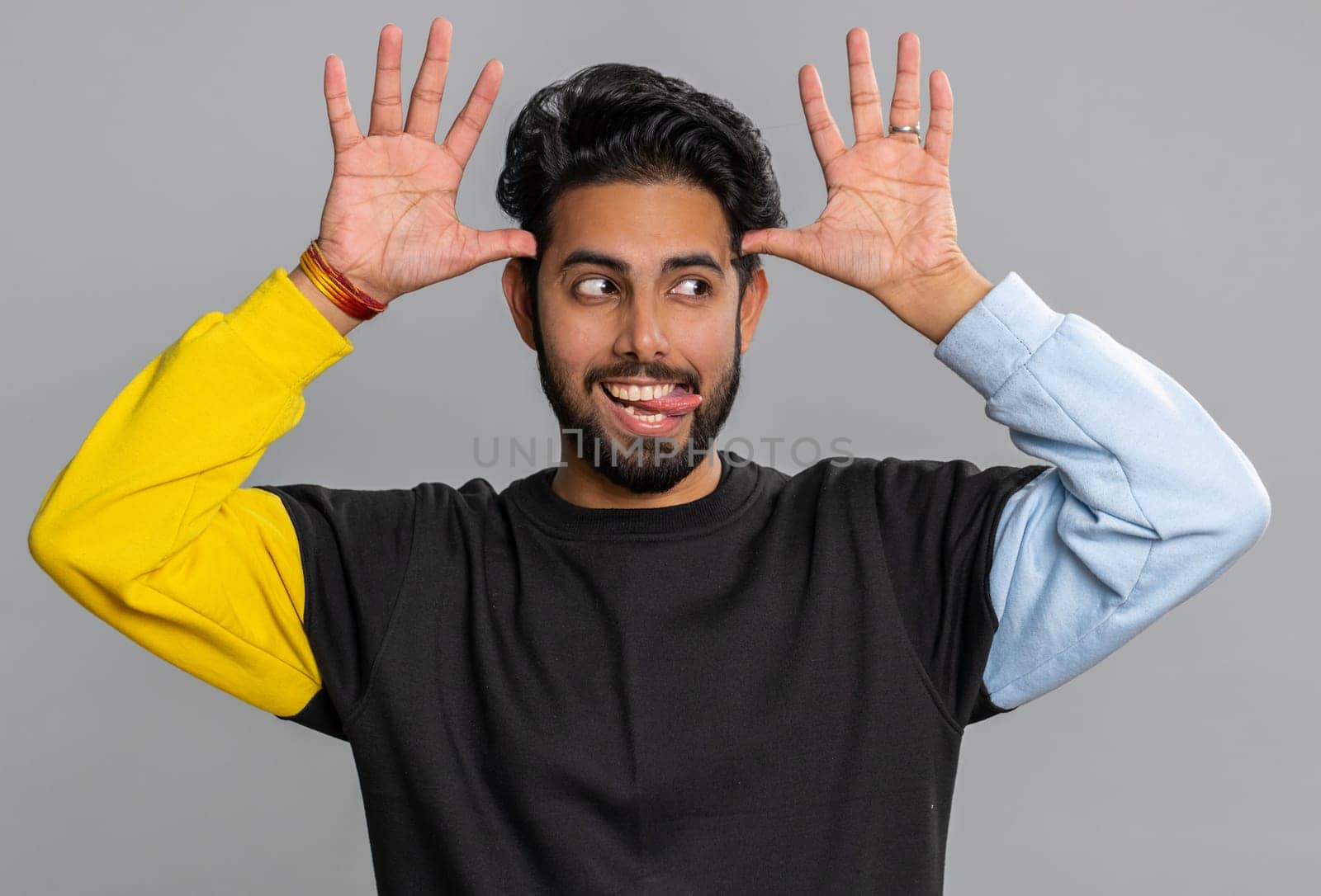 Comical funny young indian man making playful silly facial expressions and grimacing, fooling around, showing tongue. Handsome hindu crazy guy bullying isolated on studio gray background indoors