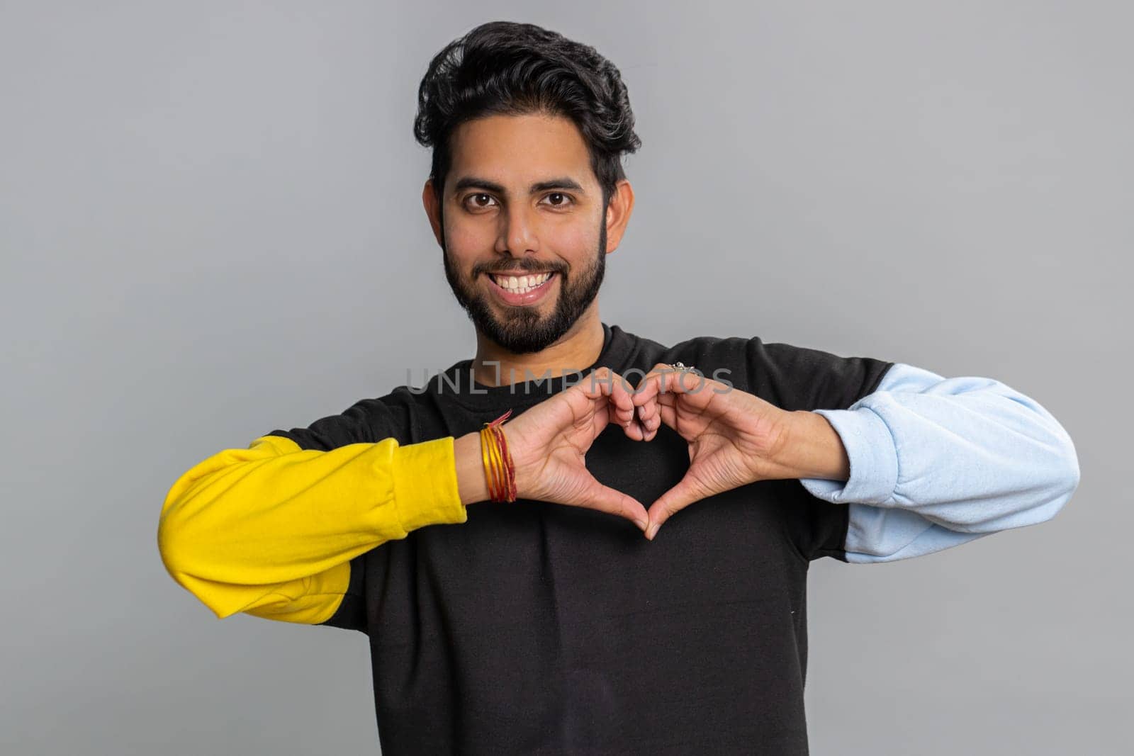 Man in love. Smiling bearded indian man 25 years old makes heart gesture demonstrates love sign expresses good feelings and sympathy. Handsome hindu young guy isolated alone on gray studio background