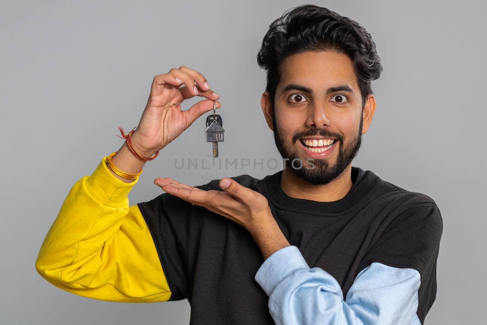 Young indian man real estate agent lifting hand showing the keys of new home house apartment, buying or renting property, mortgage loan. Bearded hindu guy isolated on gray studio background, indoors