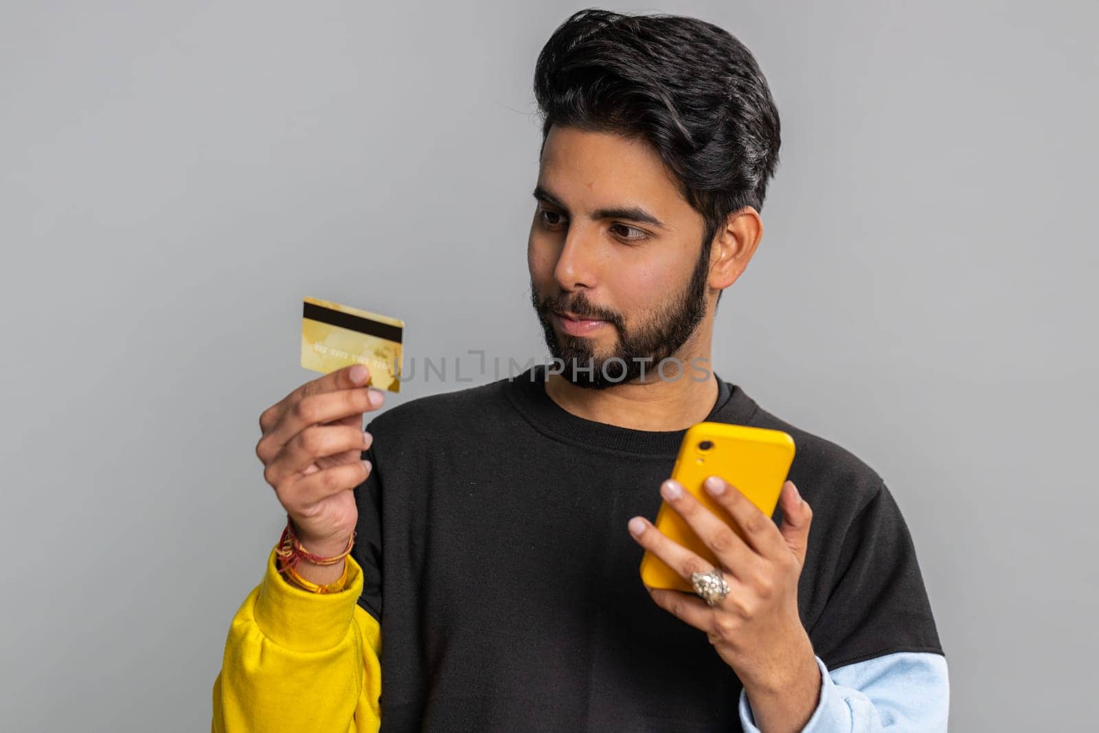 Sincere indian man customer using credit bank card and smartphone while transferring money, purchases online shopping, payment. Finance and internet. Handsome guy isolated alone on gray background