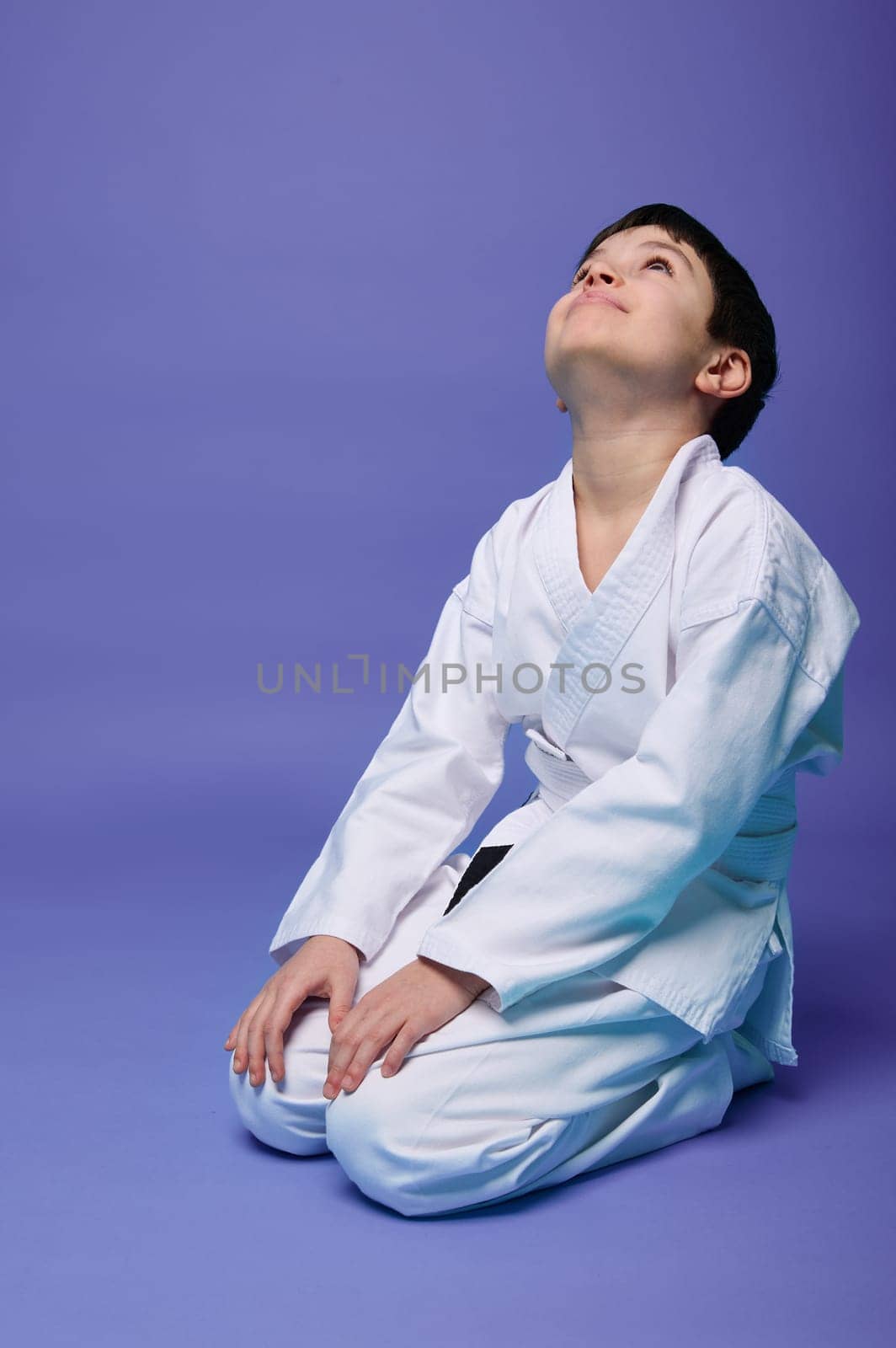 Charming pleasant Caucasian teenage boy in white kimono practicing oriental martial arts, isolated on violet background with copy ad space for text