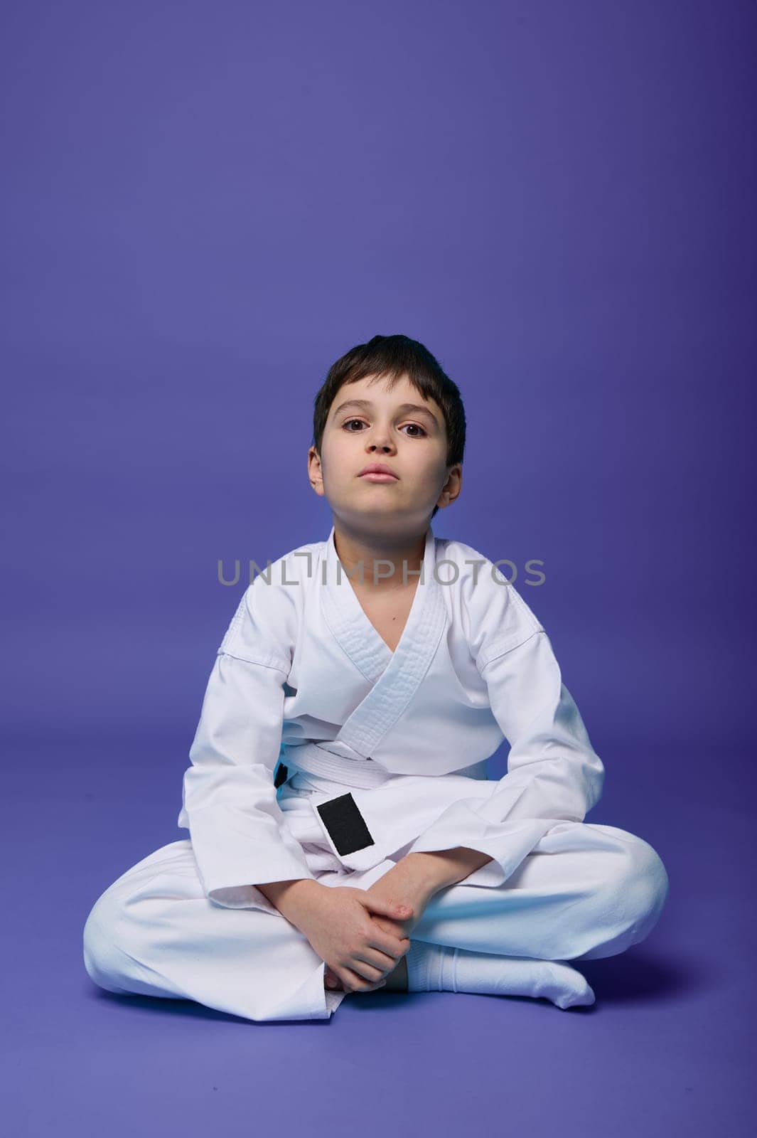 Charming confident Caucasian teenager - aikido fighter - in white kimono sitting in lotus position while practicing oriental martial arts on purple background with copy ad space for text