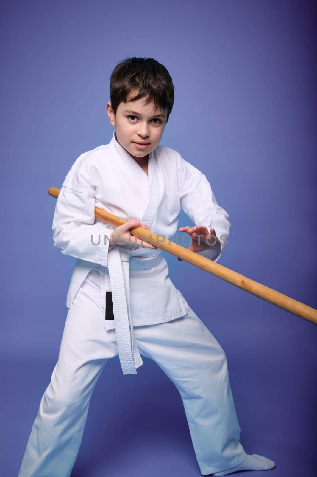 Caucasian teenage boy in a white kimono fights with a wooden sword in aikido training on a purple background with copy space for advertising text. Healthy lifestyle and martial arts concept