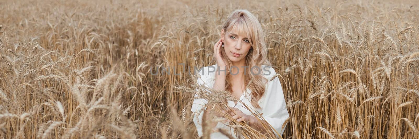 A blonde woman in a long white dress walks in a wheat field. The concept of a wedding and walking in nature.