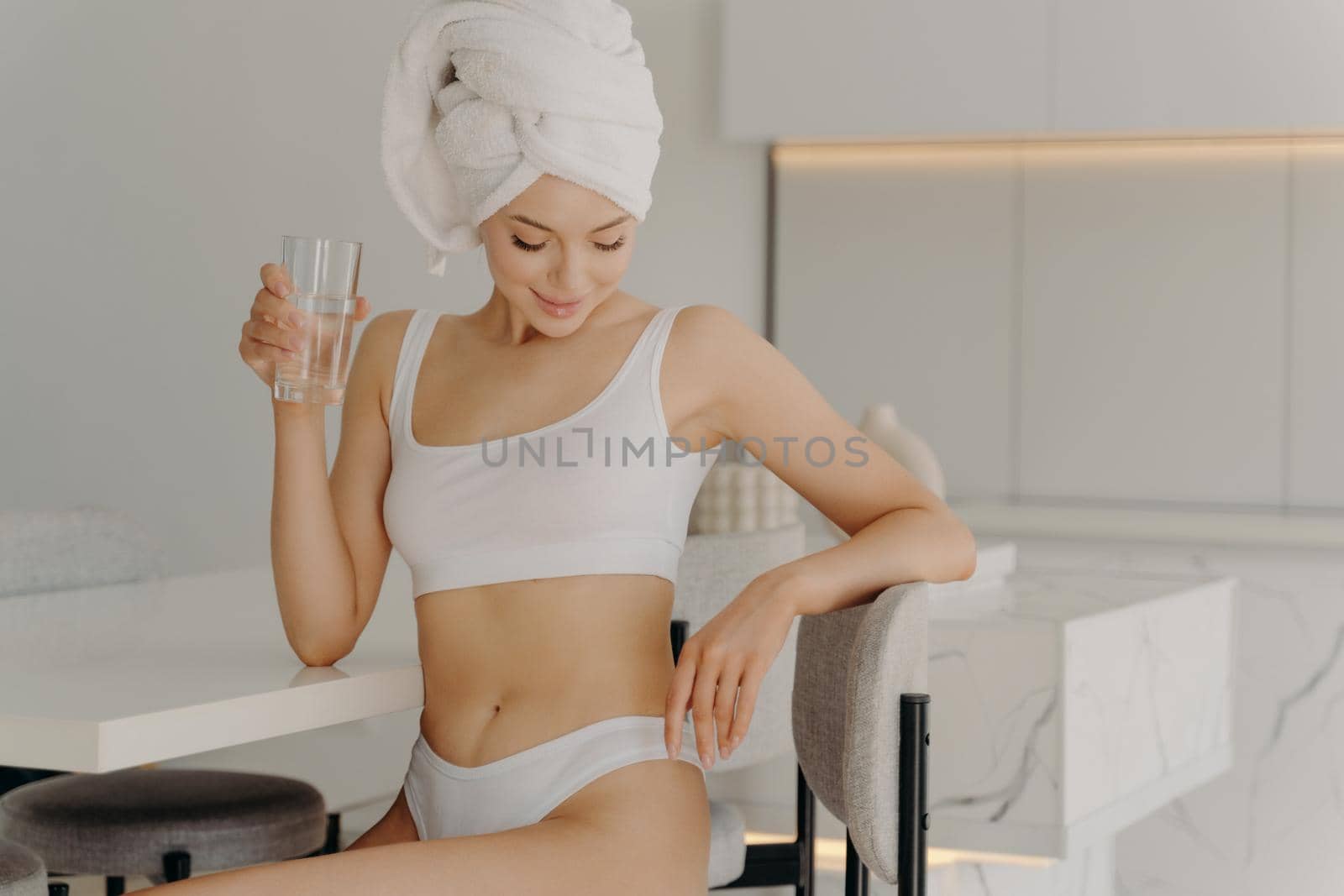 Fit attractive young woman sitting in kitchen while drinking glass of water, rehydrating herself after shower routine procedure, wears underwear and towel wrapped on head. Healthy morning habbits