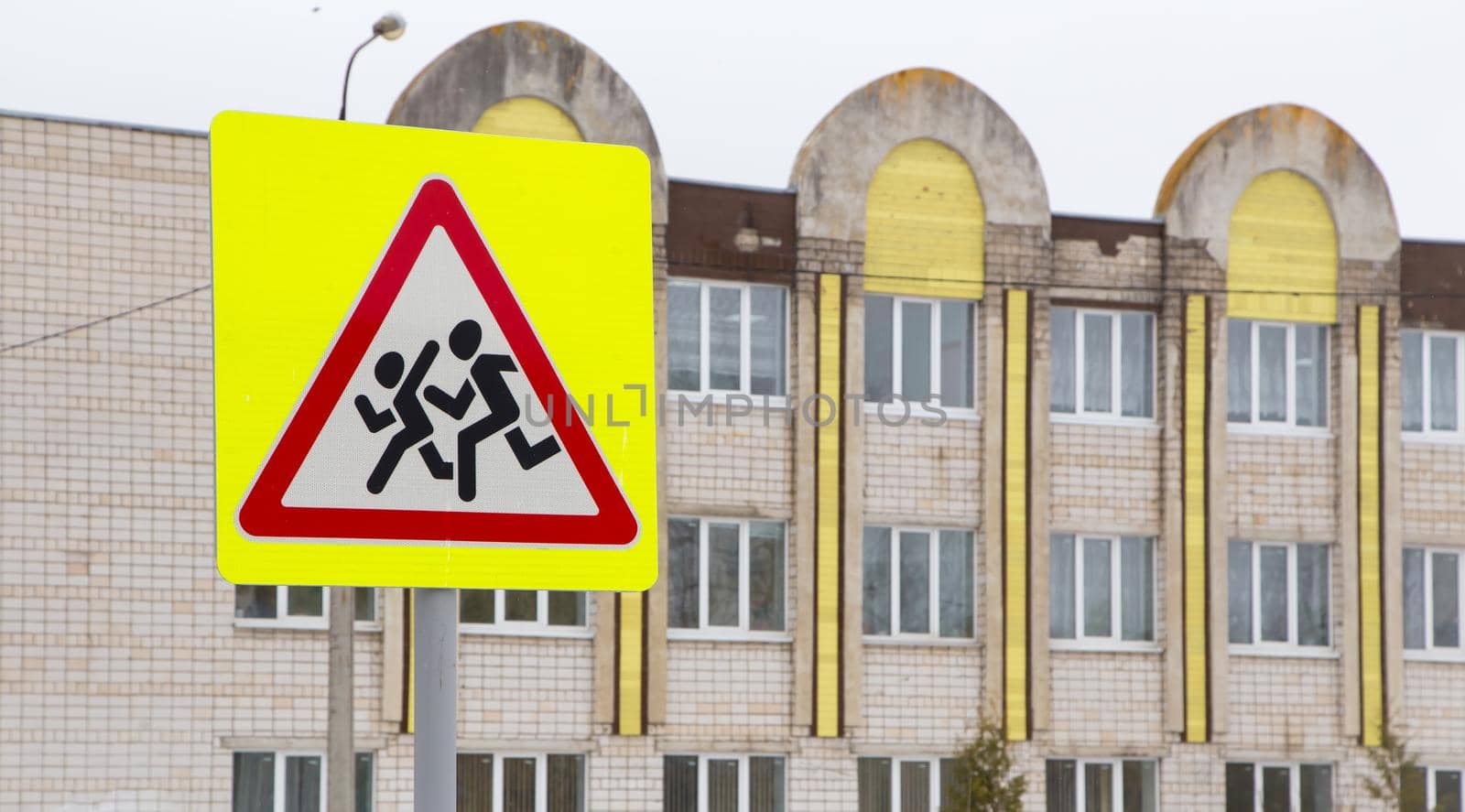 Road sign, caution children, on the background of the building. Snowdrifts with dirty snow and a large pool of melt water. Little men run on a white background in a red triangle and a yellow square.