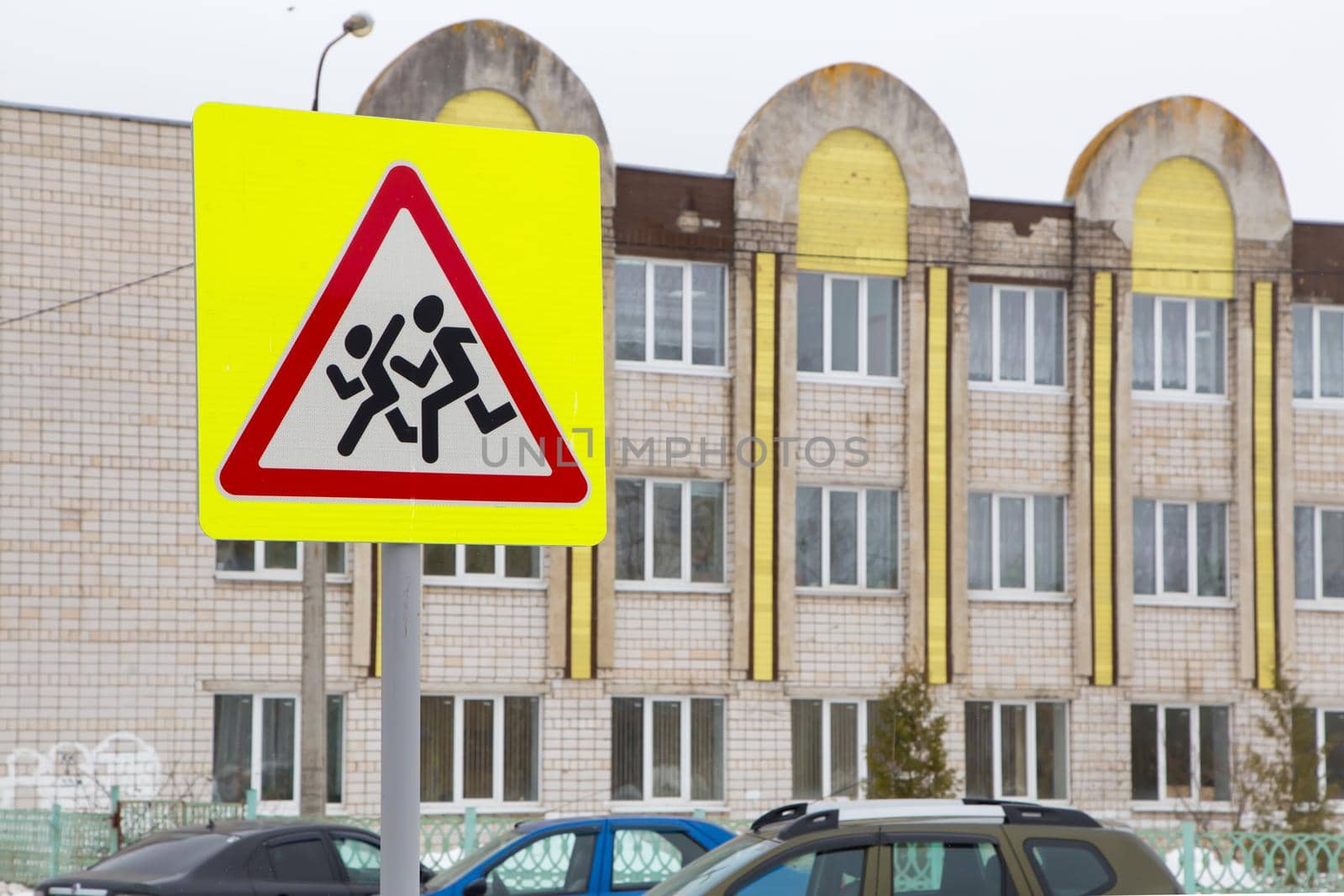 Road sign means be careful children. Against the background of the school building and cars standing nearby. Humans run on a white background in a red triangle and a yellow square. Warning sign.