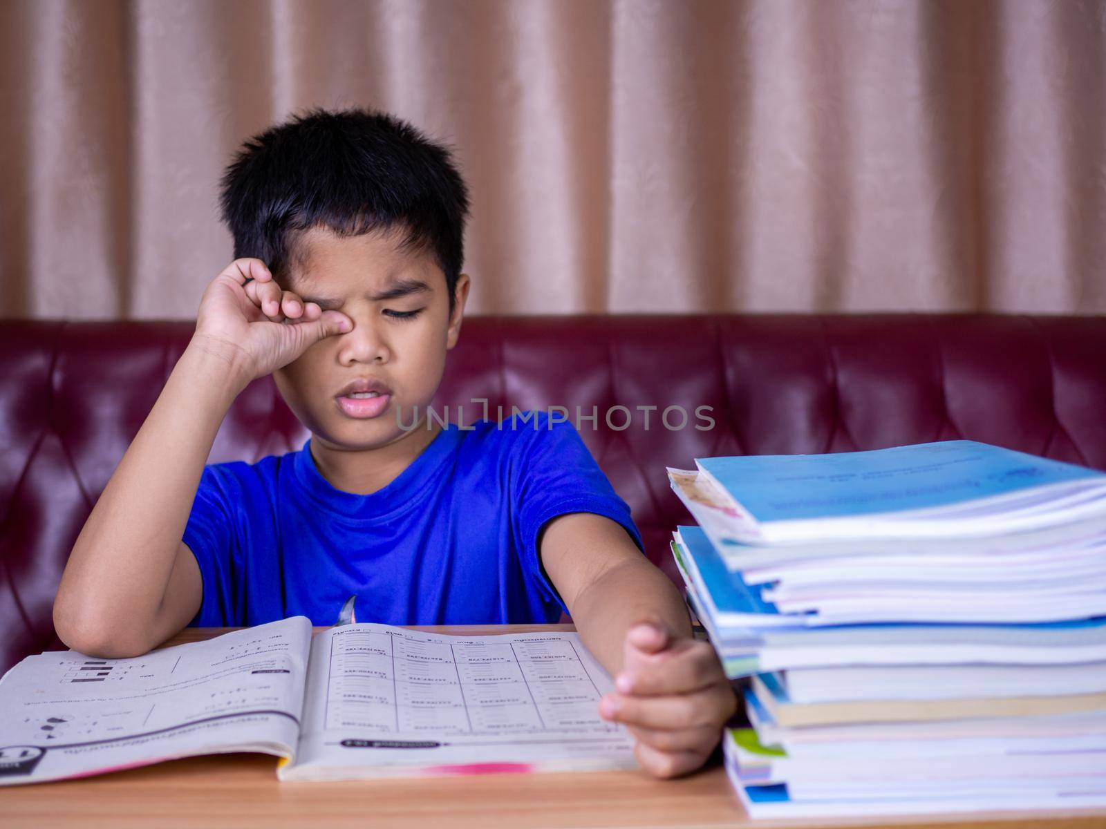 A boy is tired of reading a book on a wooden table. with a pile of books beside The background is a red sofa and cream curtains. by Unimages2527