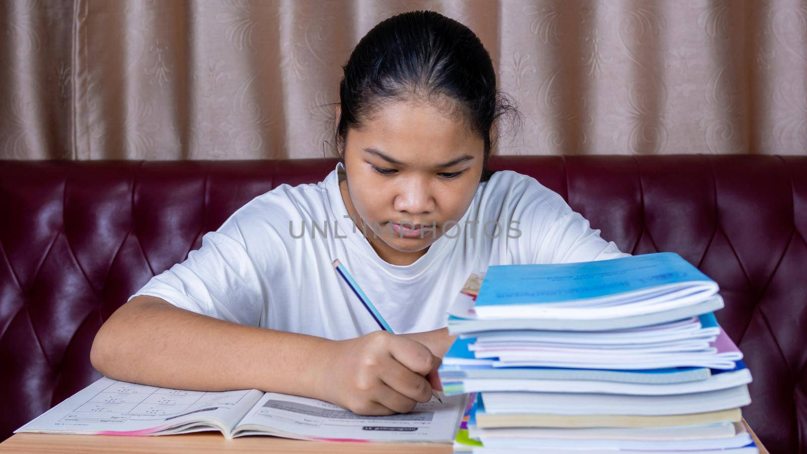 girl doing homework on a wooden table and there was a pile of books next to it The background is a red sofa and cream curtains. by Unimages2527