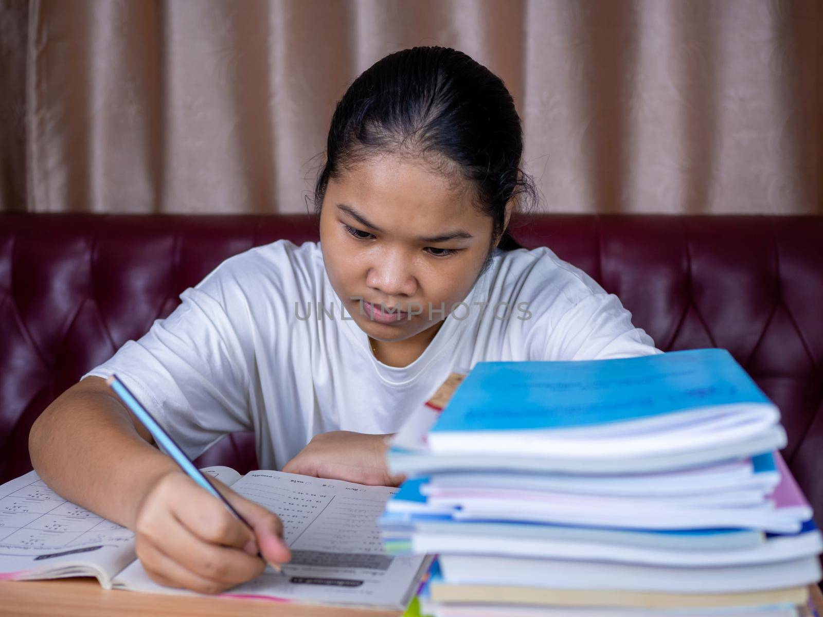 girl doing homework on a wooden table and there was a pile of books next to it The background is a red sofa and cream curtains. by Unimages2527