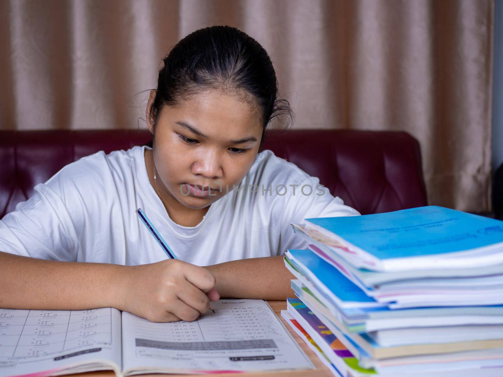 girl doing homework on a wooden table and there was a pile of books next to it The background is a red sofa and cream curtains. by Unimages2527