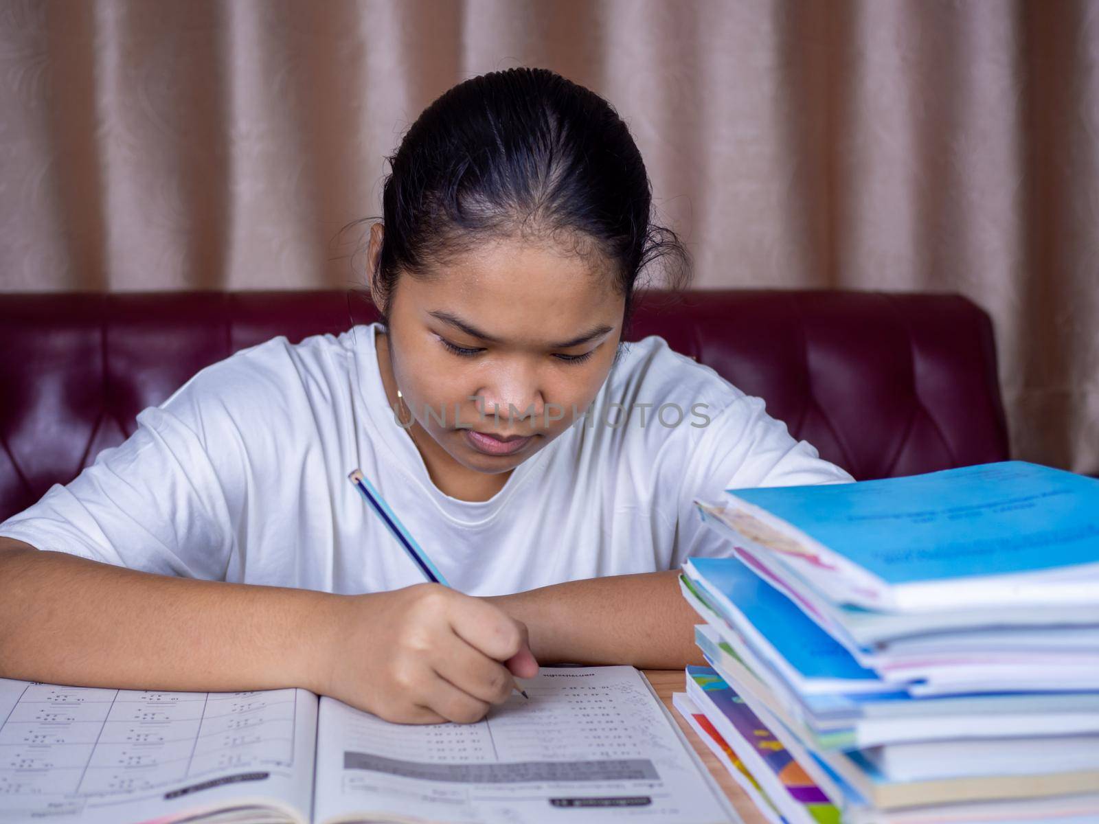 girl doing homework on a wooden table and there was a pile of books next to it The background is a red sofa and cream curtains. by Unimages2527