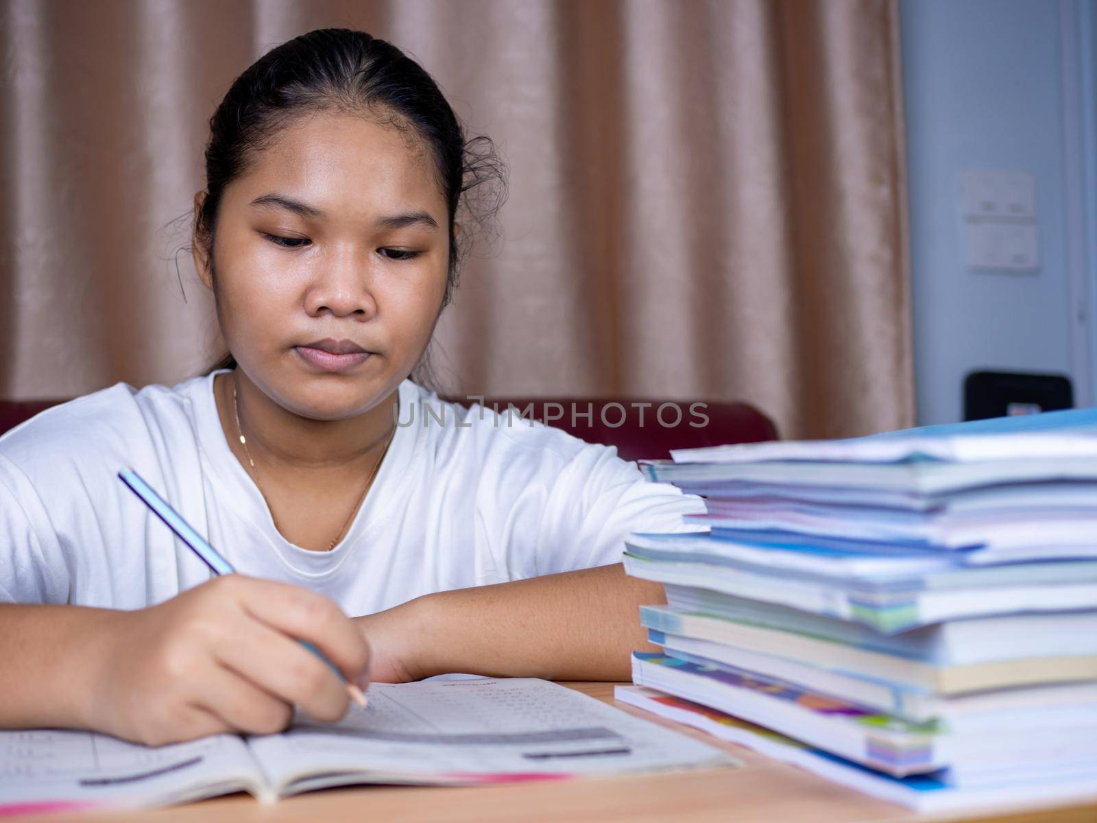 girl doing homework on a wooden table and there was a pile of books next to it The background is a red sofa and cream curtains. by Unimages2527