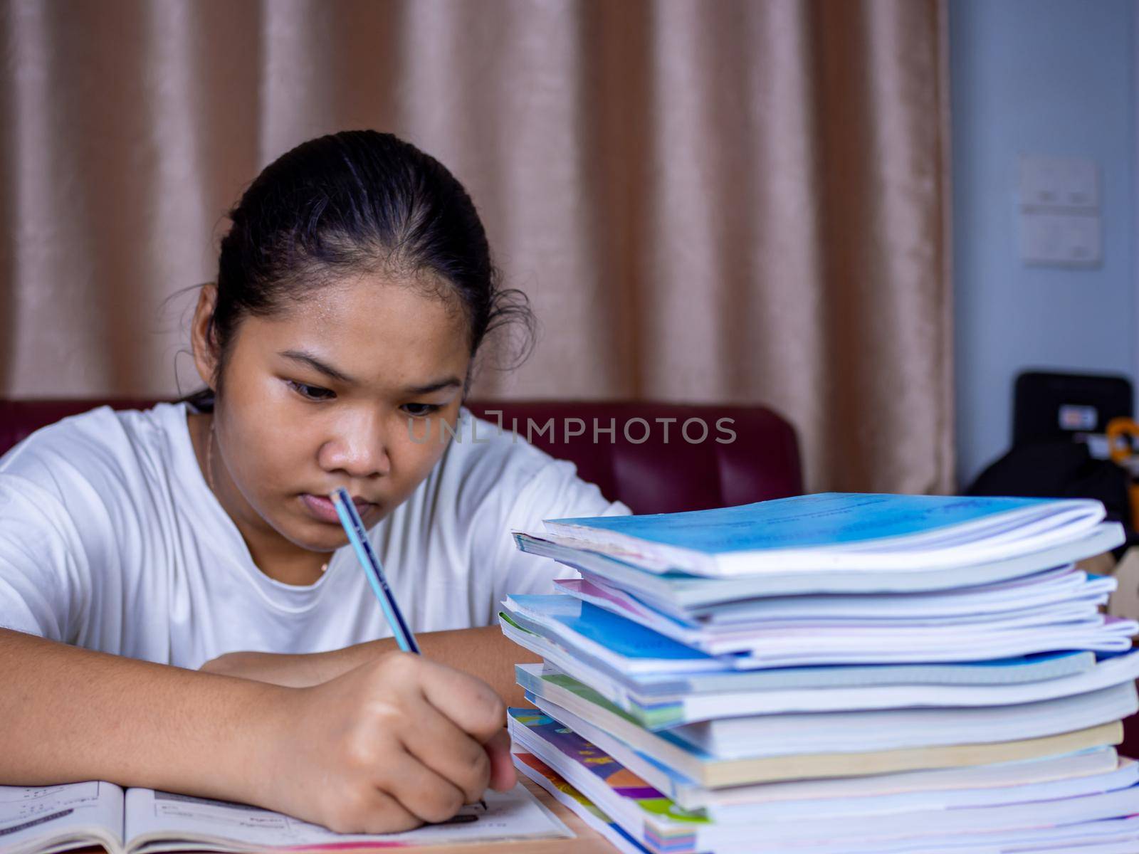 girl doing homework on a wooden table and there was a pile of books next to it The background is a red sofa and cream curtains.