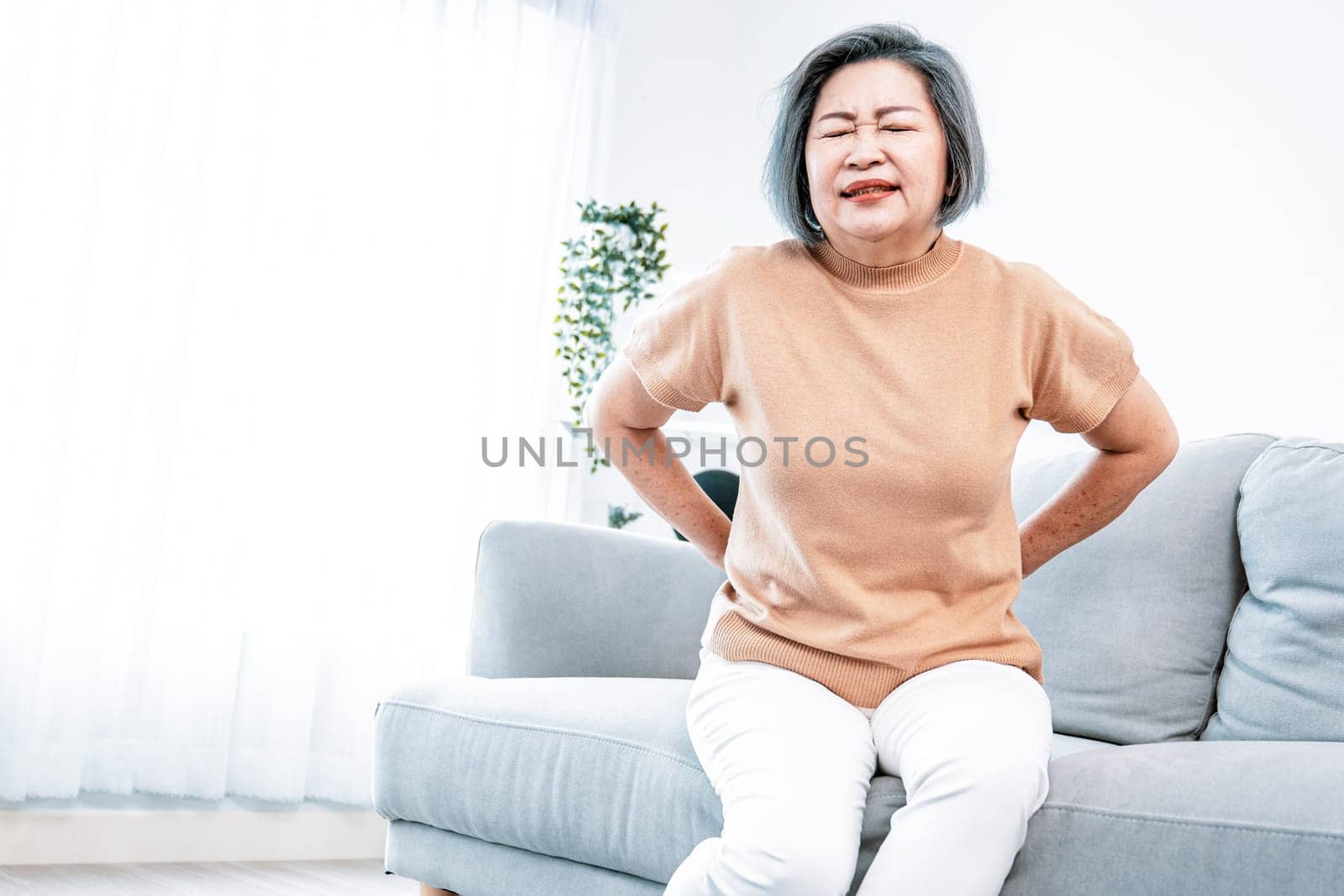 An agonizing senior woman experiencing back pain while sitting on her sofa at home and holding her back.