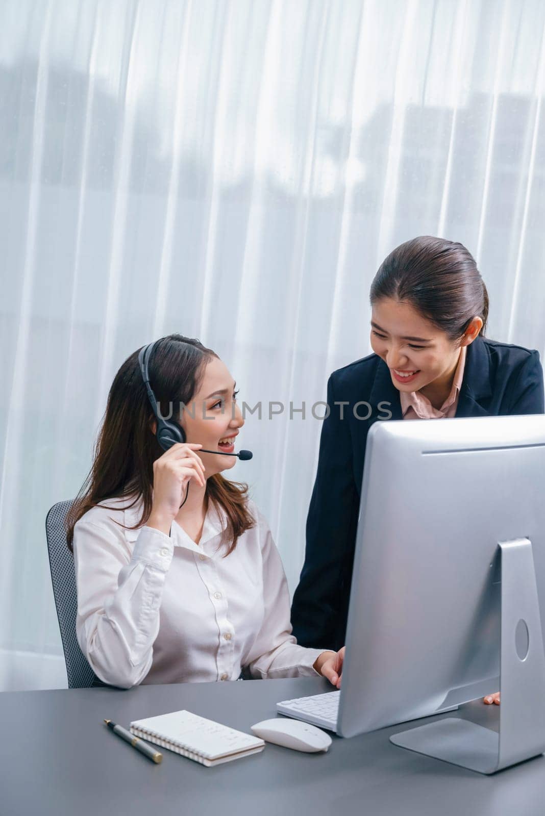 Asian female customer support operator wearing headset is guided by her supportive manager. Experienced colleague help operator handle a call with a client, providing advice and guidance. Enthusiastic