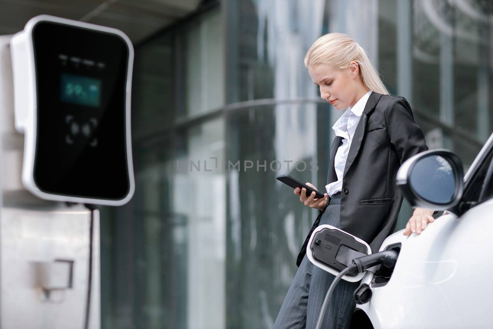 Progressive businesswoman with mockup smartphone with EV at charging station. by biancoblue