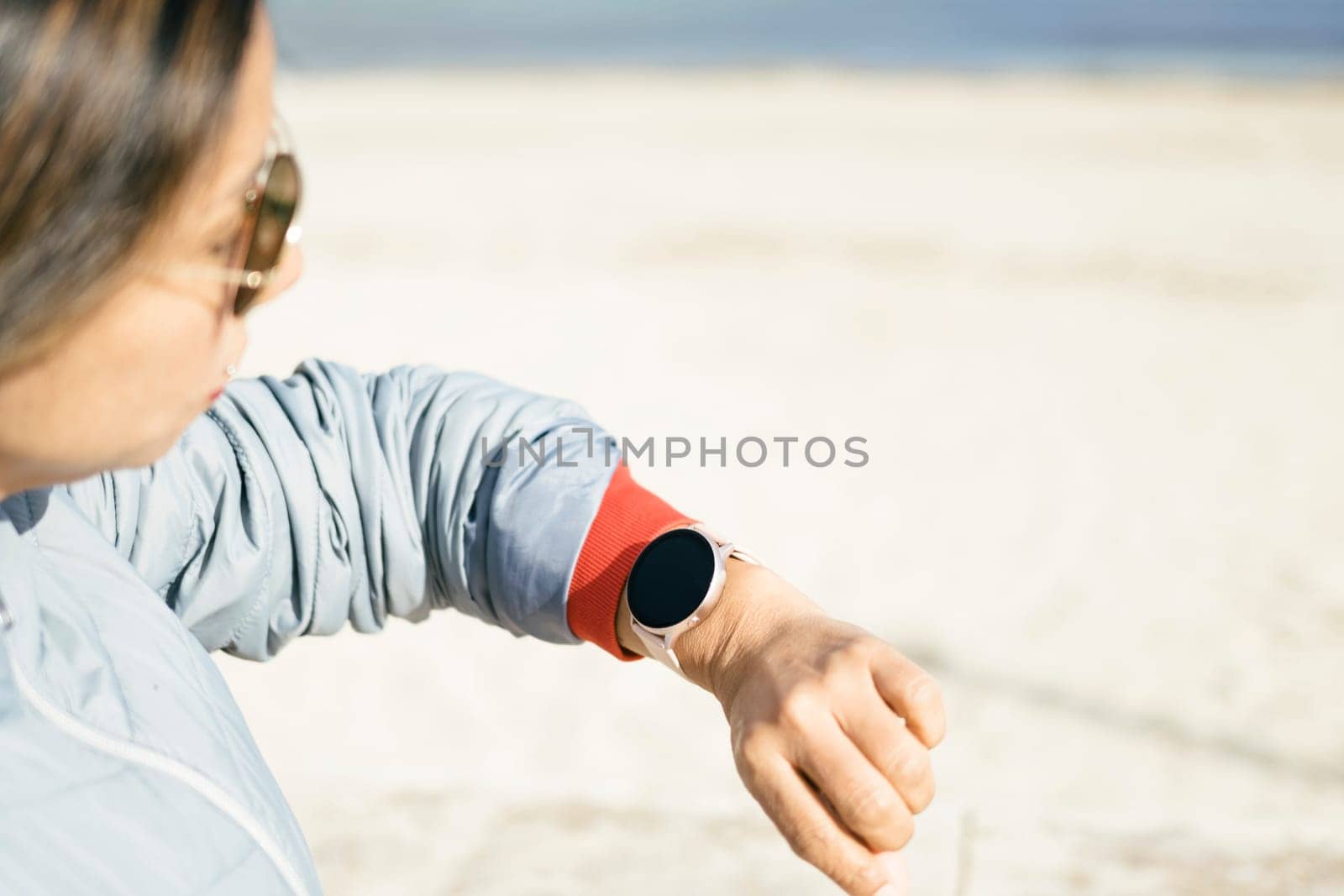 view of latina woman looking at smart watch, with sunglasses blue jacket on a sunny day with light background to copy space on the right