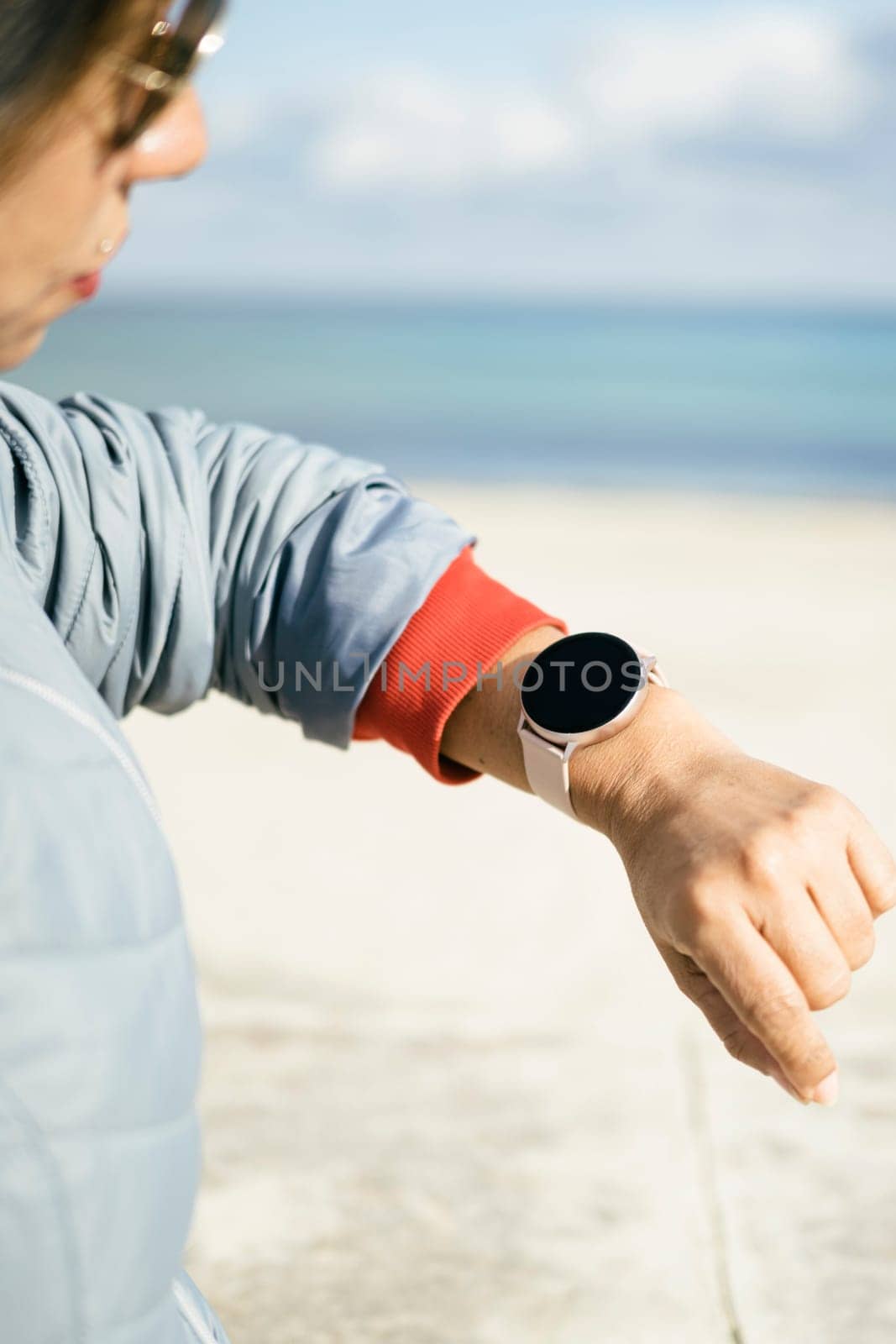 Cropped photo of a stylish woman checks notification on wearable digital wrist watch monitors the weather wears casual jacket poses on the street. Woman uses modern gadget on the street