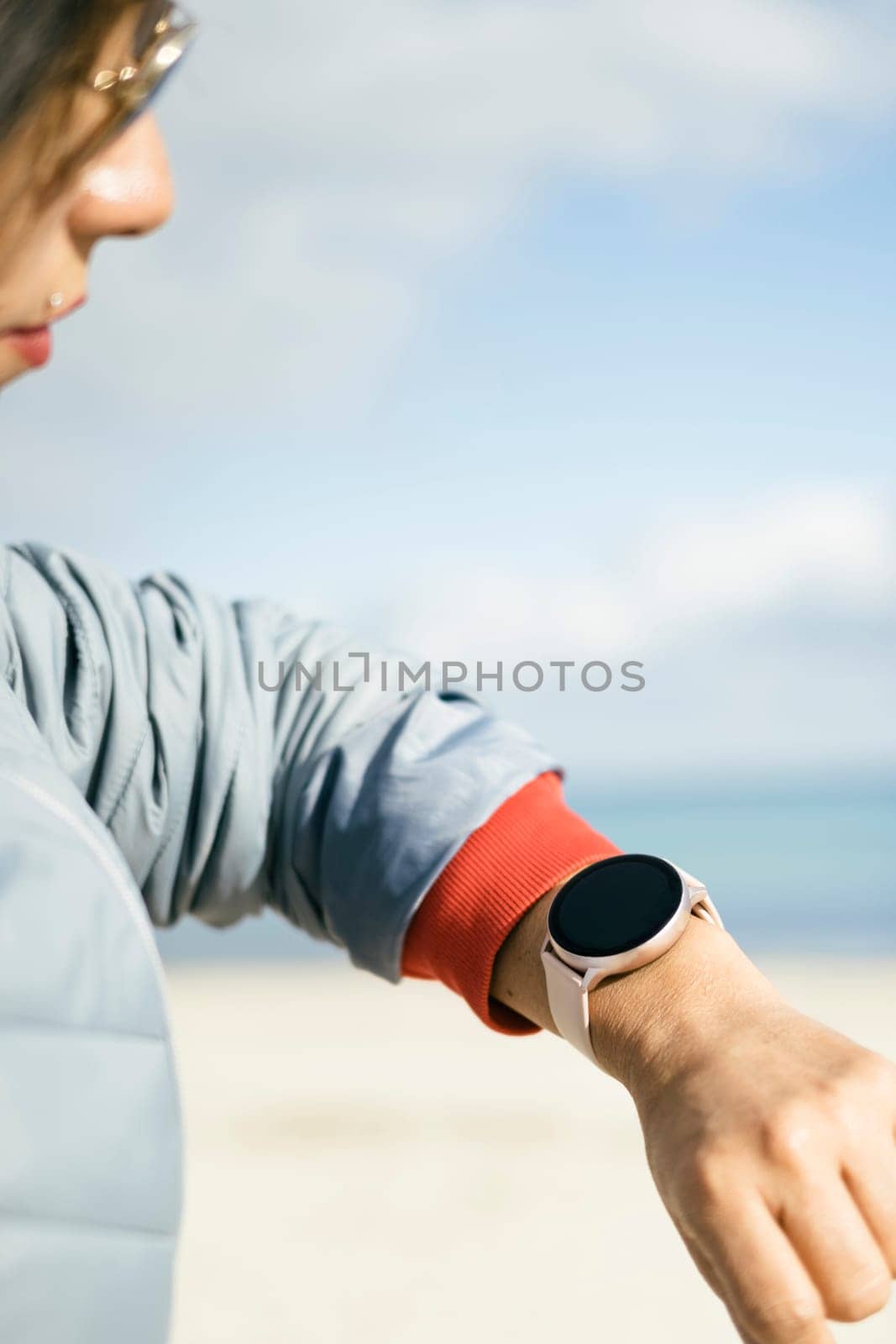 close-up view of latina woman looking at smart watch, wearing sunglasses blue jacket on a sunny day with clear background of a beach and blue sky to copy space to above