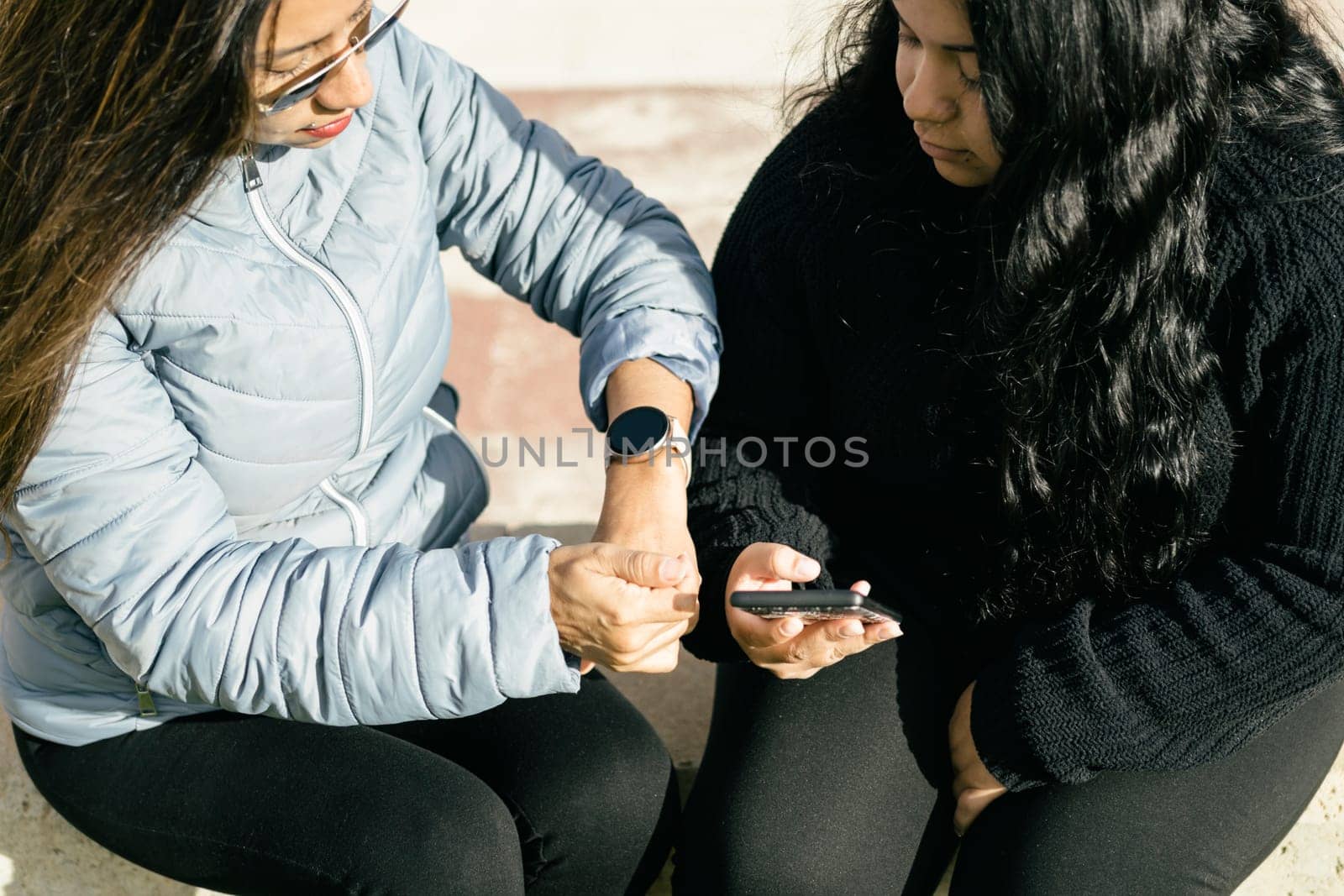 This stock photo features two Latin women sitting together and sharing data with smart electronic devices. Both women are looking at their respective devices, which are displaying information on their screens. One woman is holding a smartphone while the other has a tablet in front of her. The two women appear to be in a casual setting, perhaps a coffee shop or office break room, as they discuss the information they are sharing. This image could be used to represent modern communication and collaboration between colleagues, friends, or family members who are using technology to share information and stay connected.