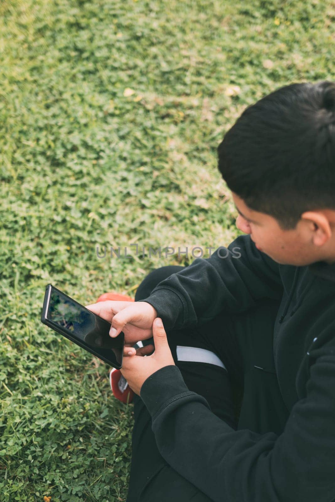 top view young millennial boy sitting in the park, in green Scutch grass with black casual clothes, looking at his smart phone, selective focus, concept of technology.