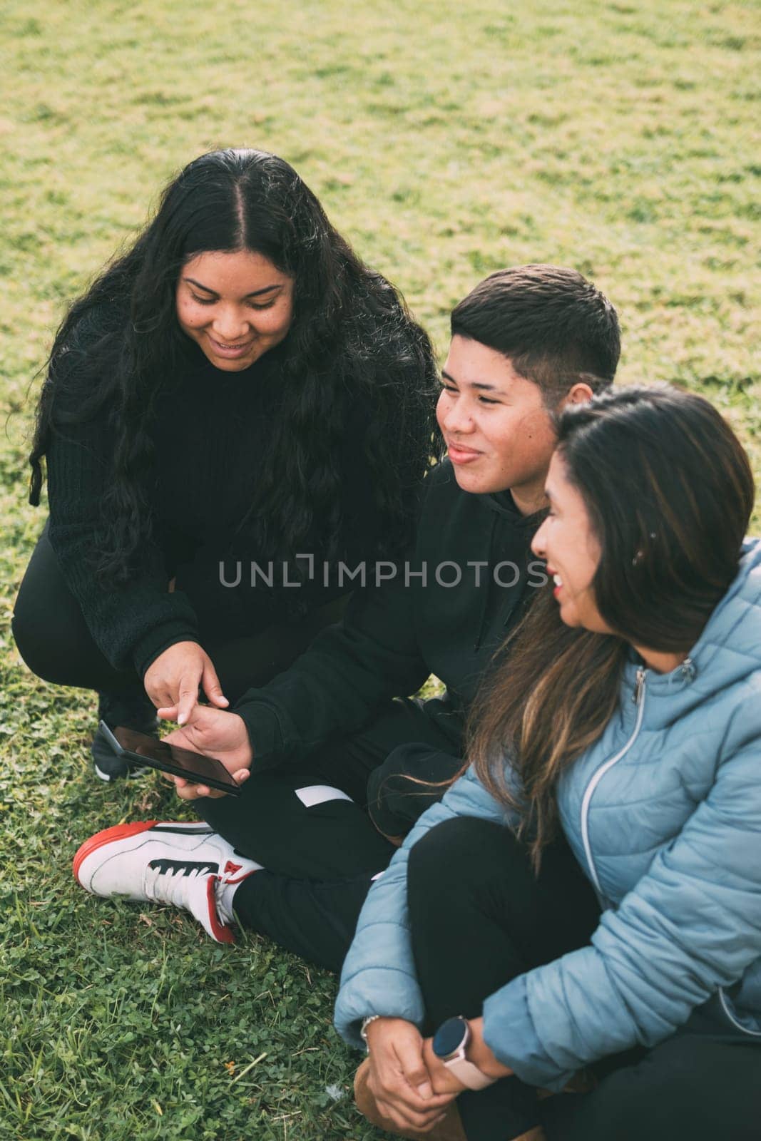 three individuals of Hispanic-Latino ethnicity sitting on the lush green grass of a park, with smart device. Relaxed and smiling expressions on their faces they are enjoying a moment of leisure amidst the hustle and bustle of everyday life.